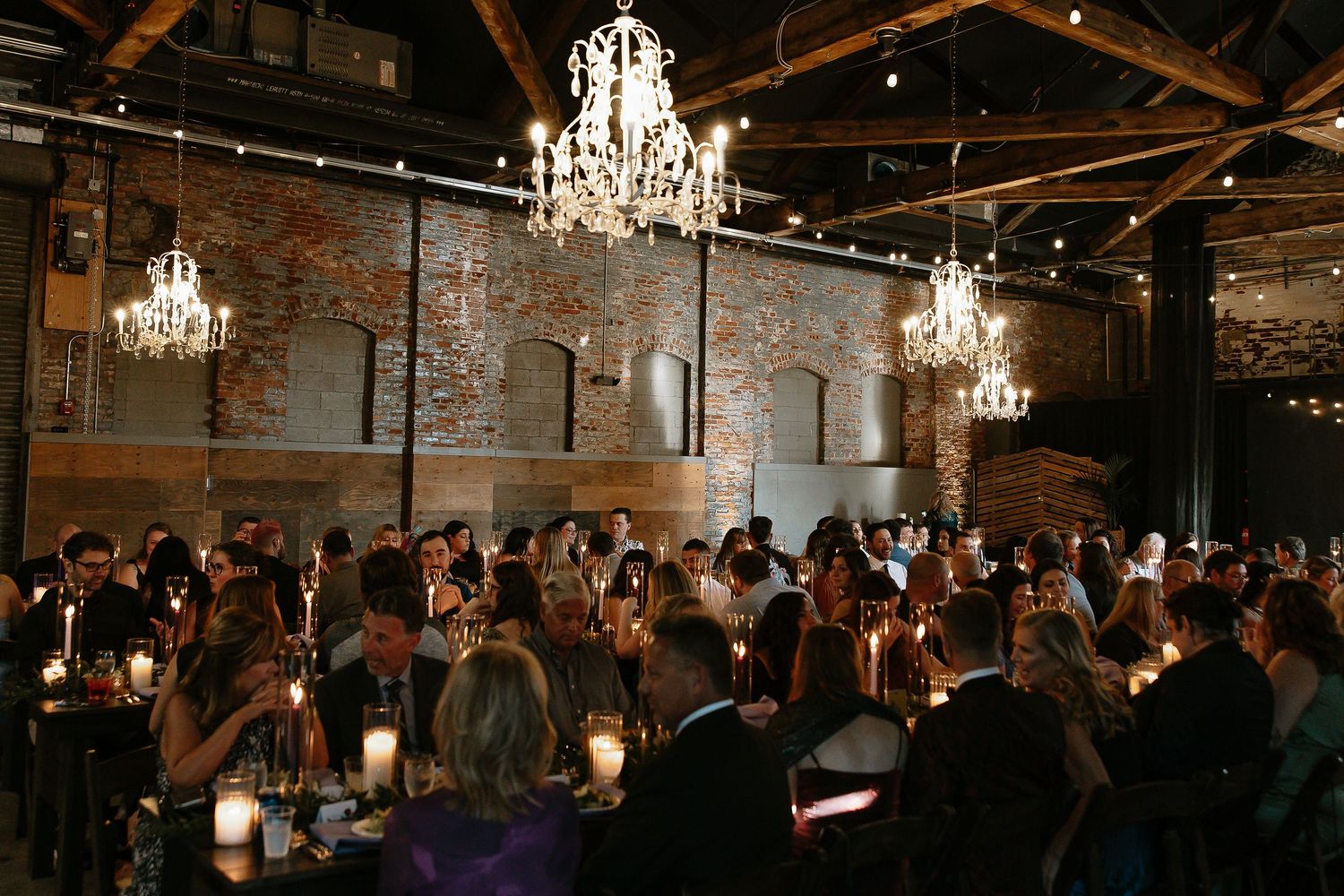 Elegant wedding reception in rustic brick venue with crystal chandeliers and candlelit tables.