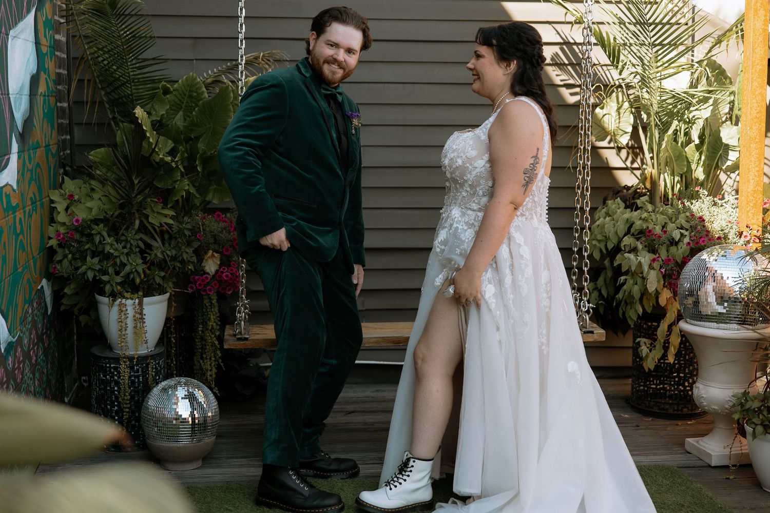 Bride in lace gown with combat boots laughs with groom in green velvet suit at outdoor wedding.