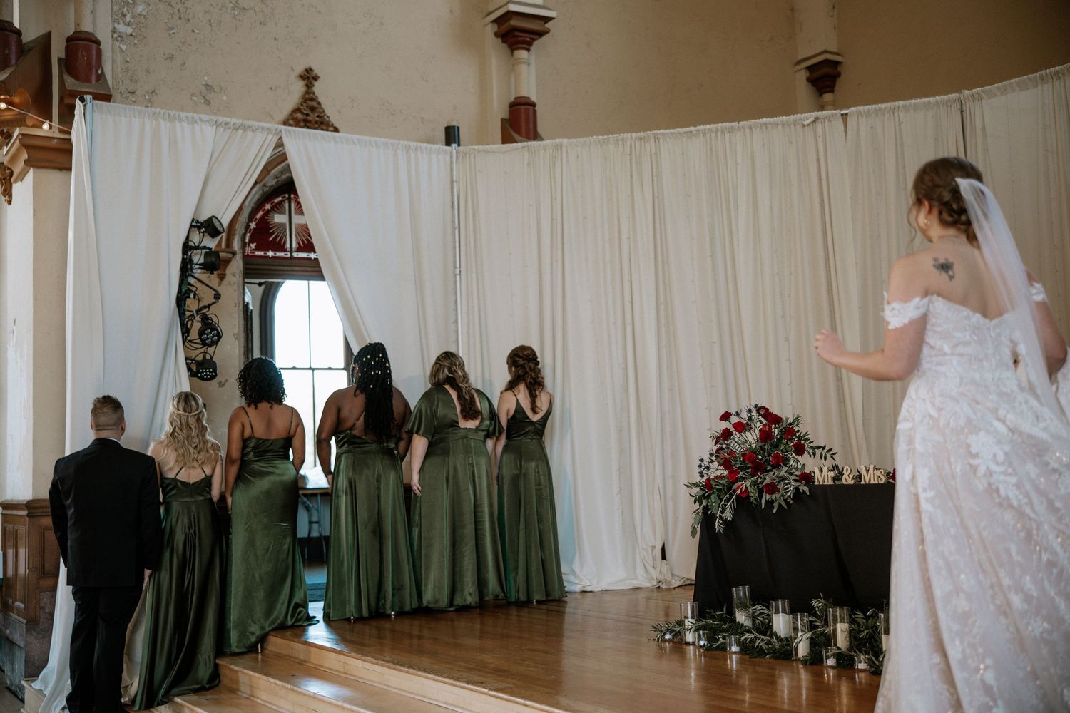 Bridesmaids in elegant olive green gowns stand at a wedding altar with white drape backdrop at Urban Artifact in Cincinnati, Ohio
