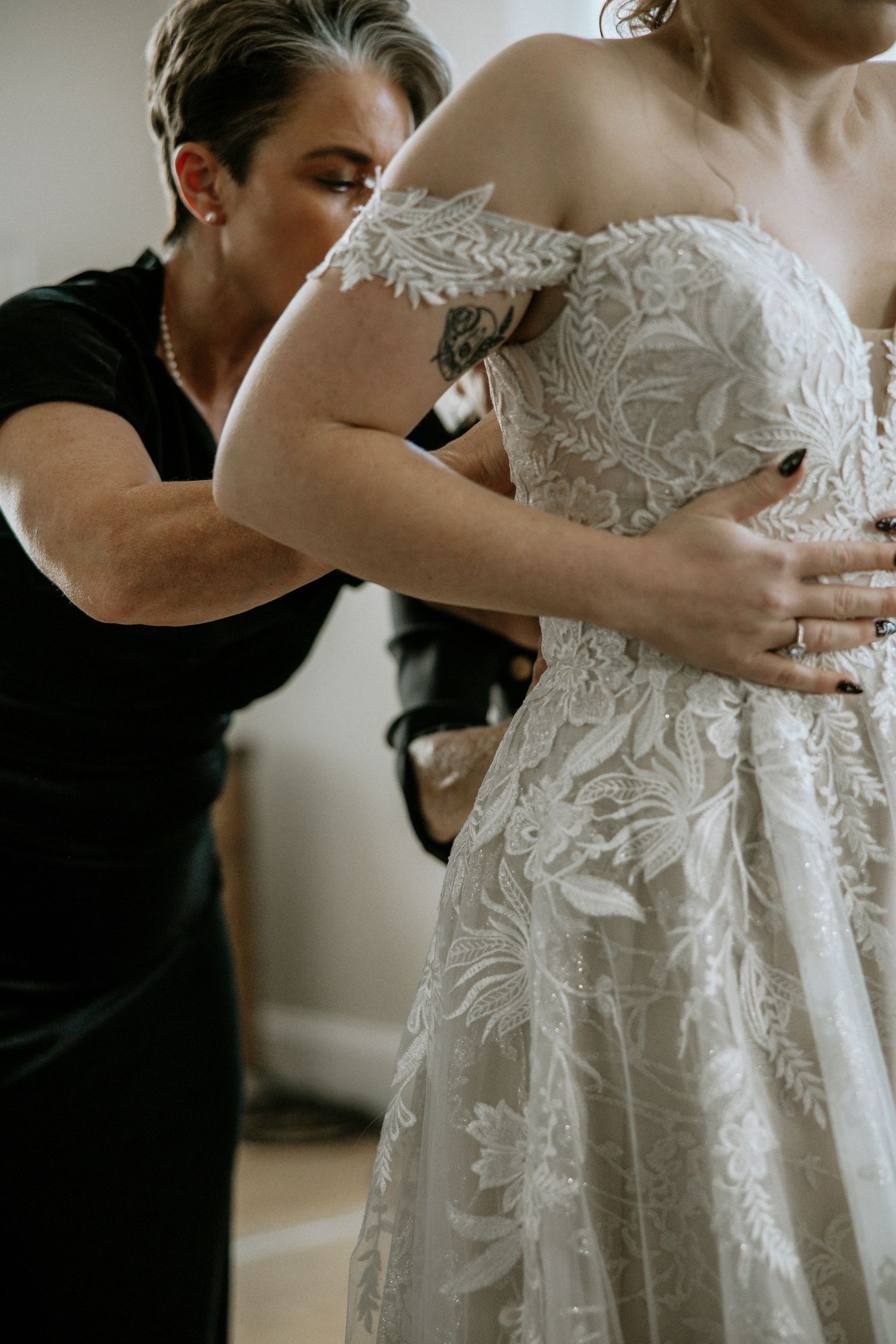 Wedding assistant helping bride into an intricate off-shoulder lace wedding gown during bridal prep at Urban Artifact in Cincinnati, Ohio