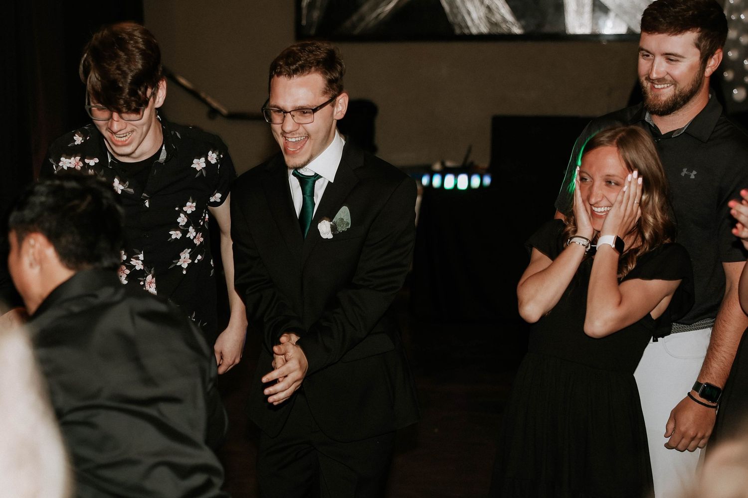 Guests dancing and laughing at a wedding reception, with a groom in a black suit and green tie celebrating on the dance floor.