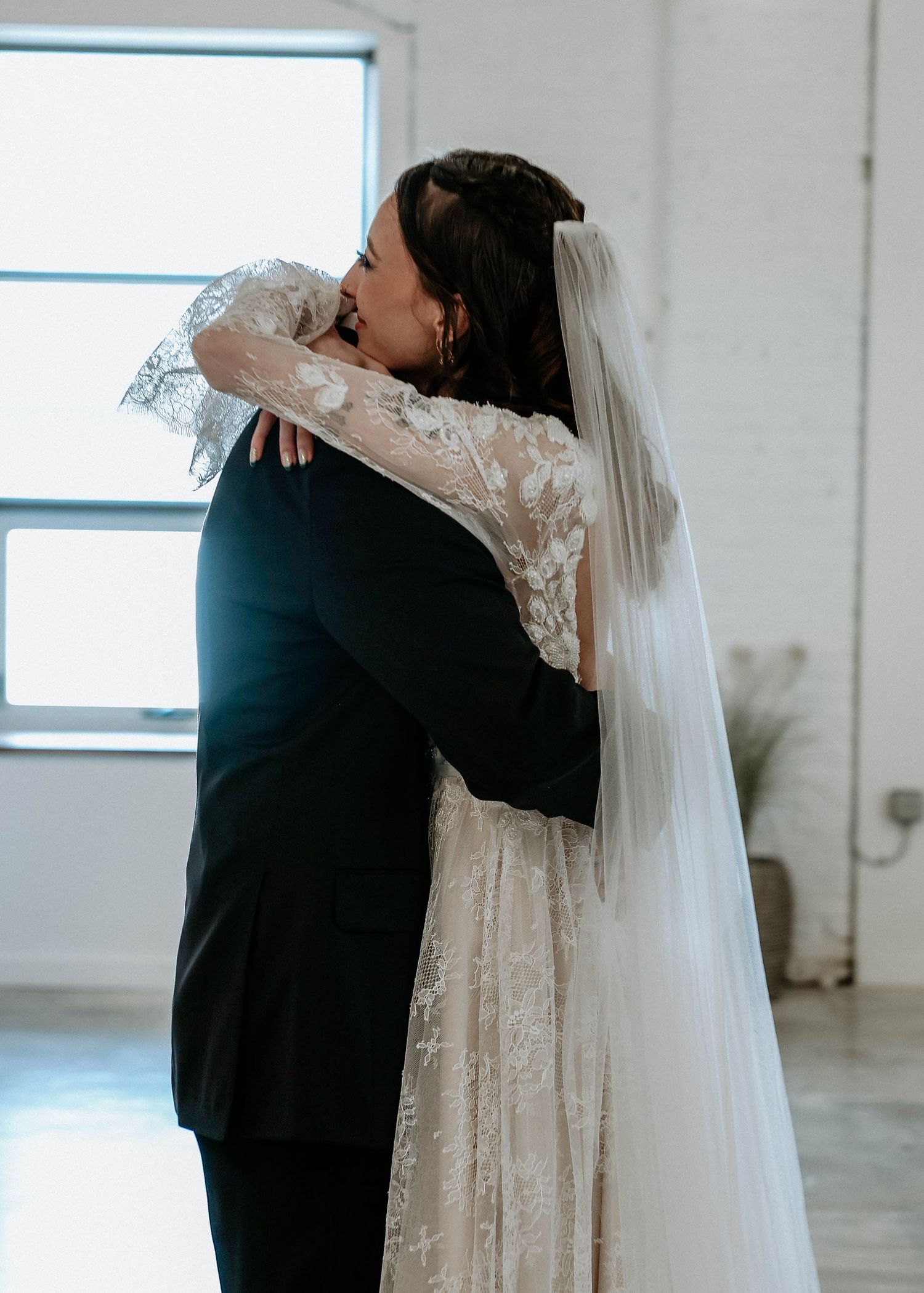 A bride in lace gown and veil embraces groom in black suit during emotional first look at indoor wedding venue.