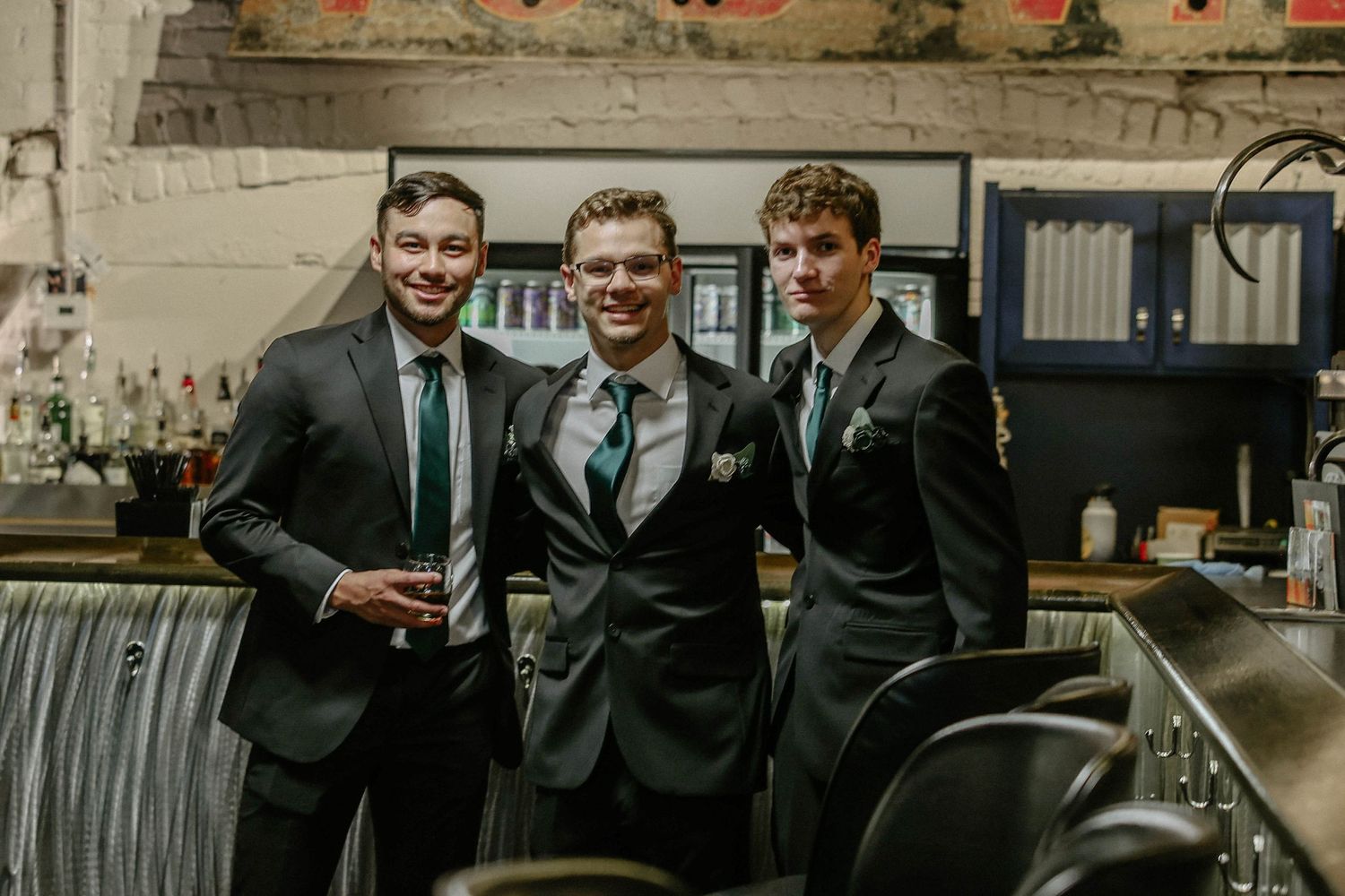 Three young men in dark suits smiling together at a bar venue during a wedding celebration.