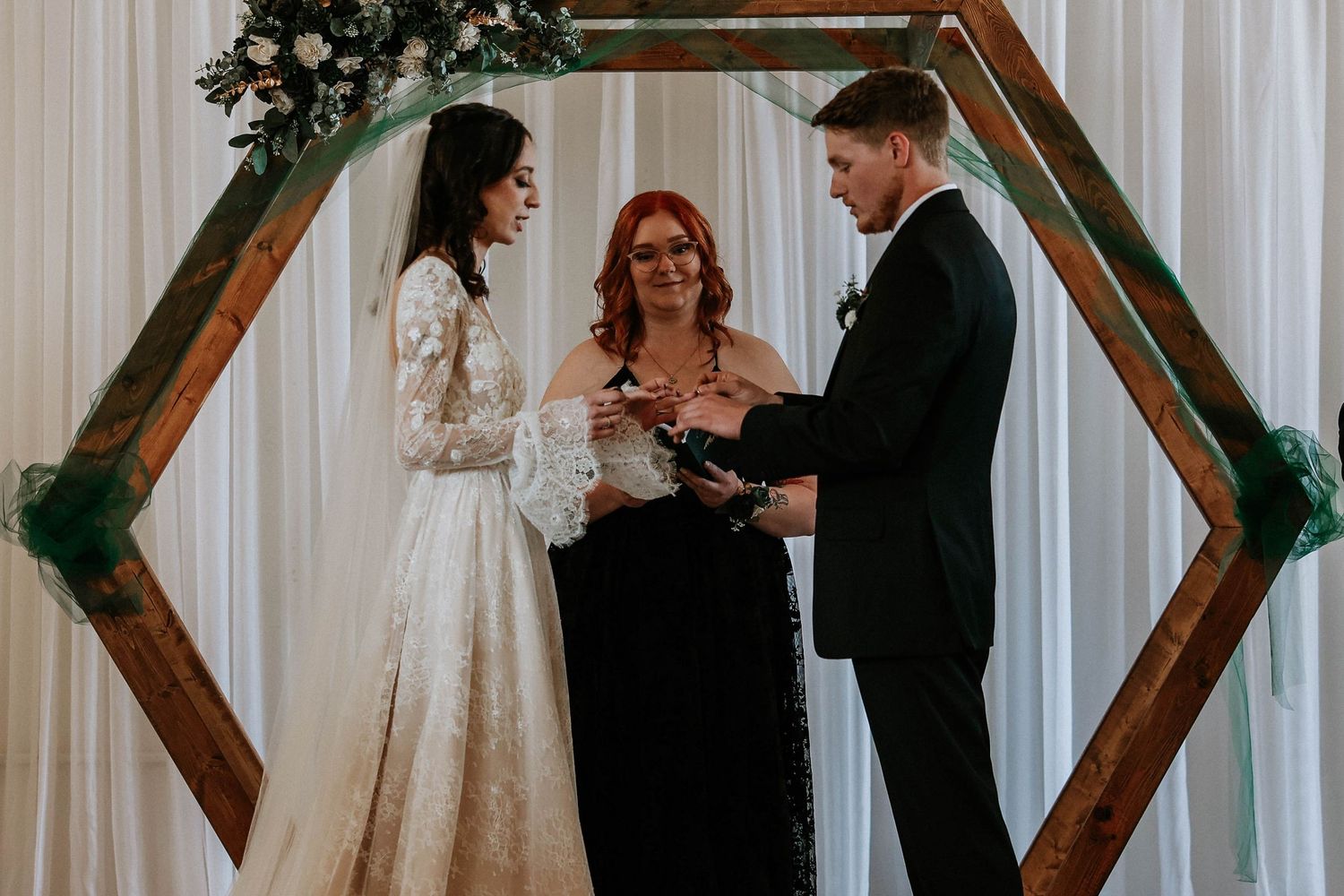 Bride and groom exchange vows under a hexagonal wooden arch with floral decor, officiated by a red-haired woman in black.