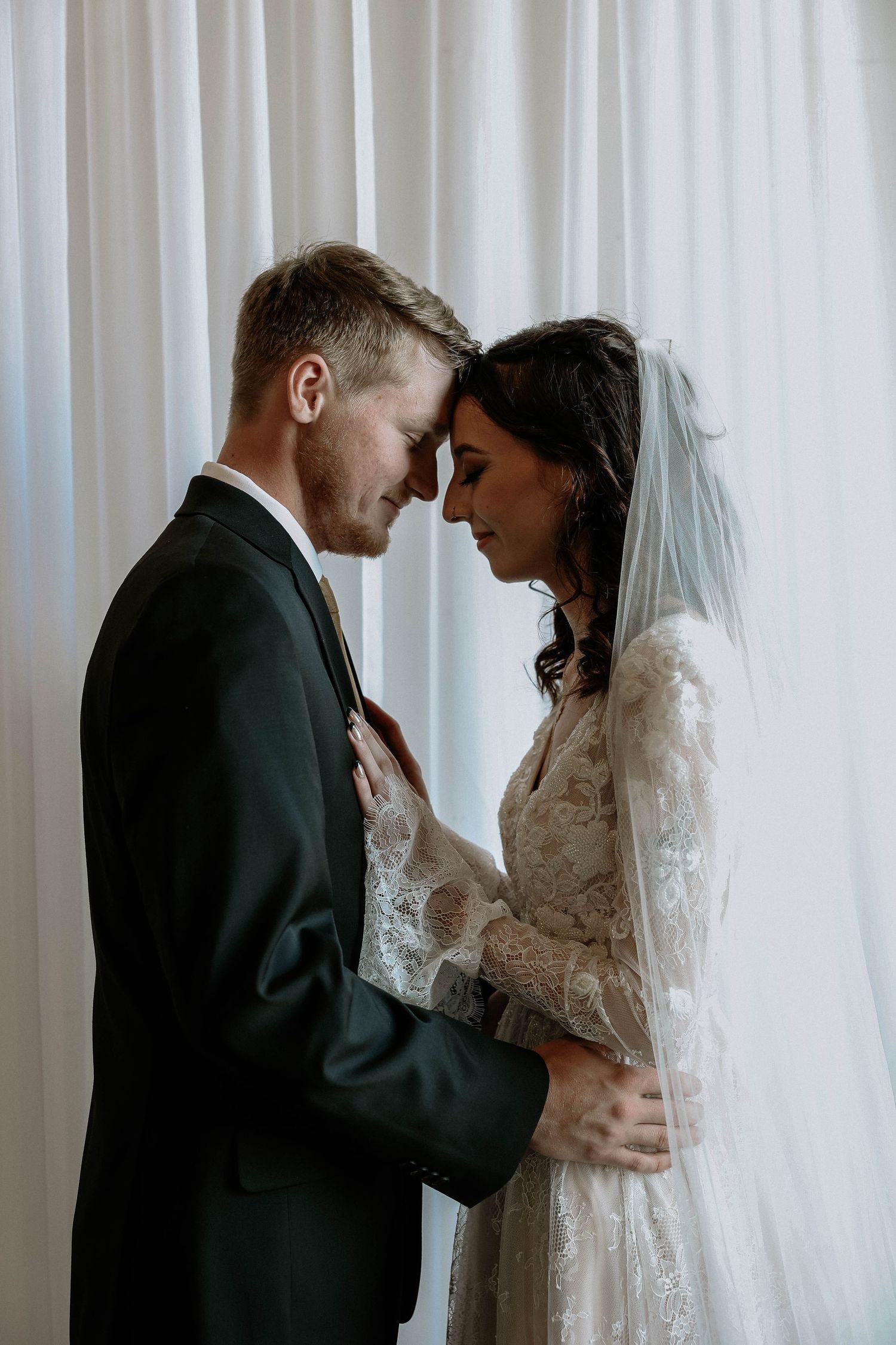 Bride and groom touching foreheads tenderly in front of white curtains, wearing lace wedding dress and black suit.