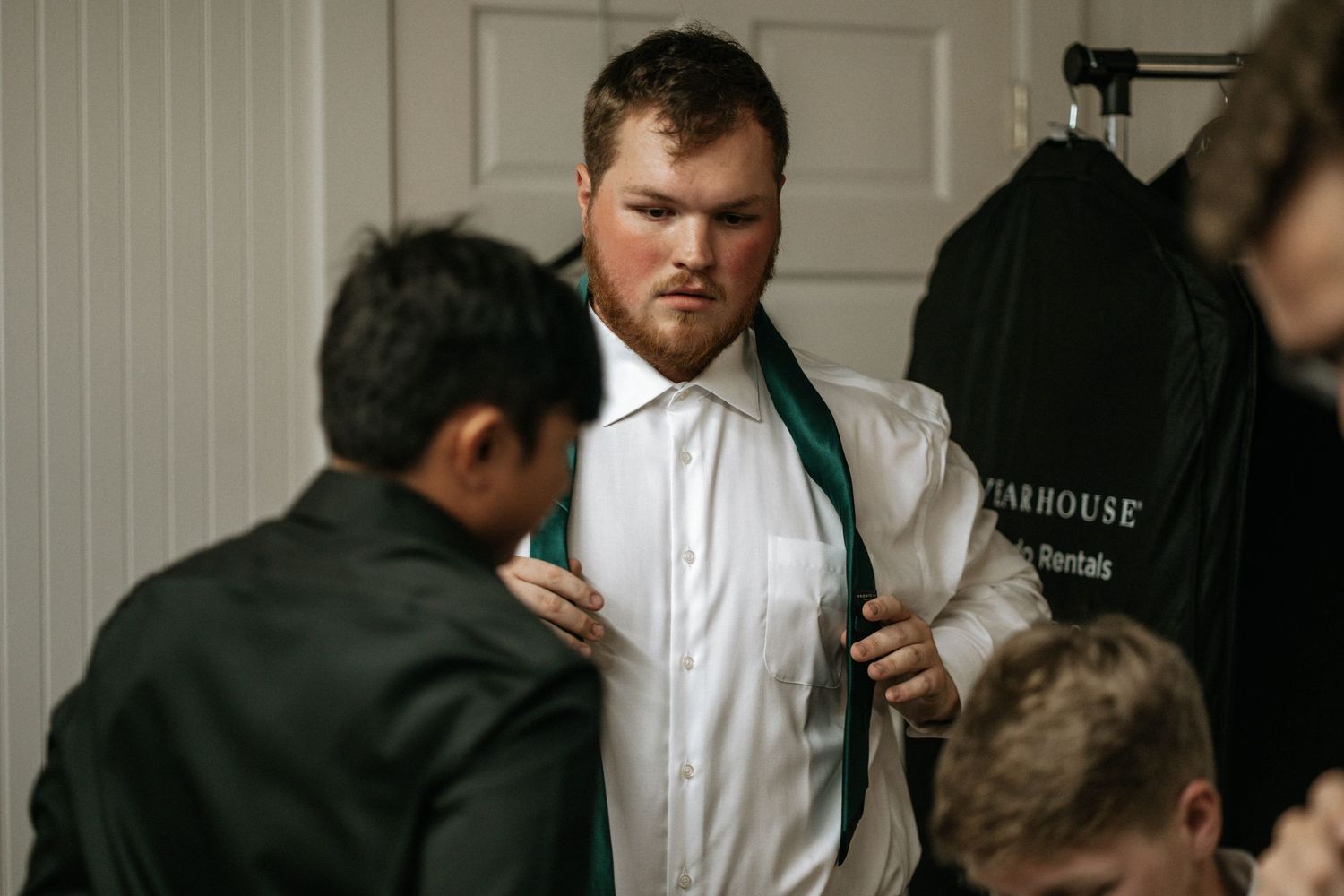 Man in white dress shirt being helped with a green tie while getting dressed for a formal event.