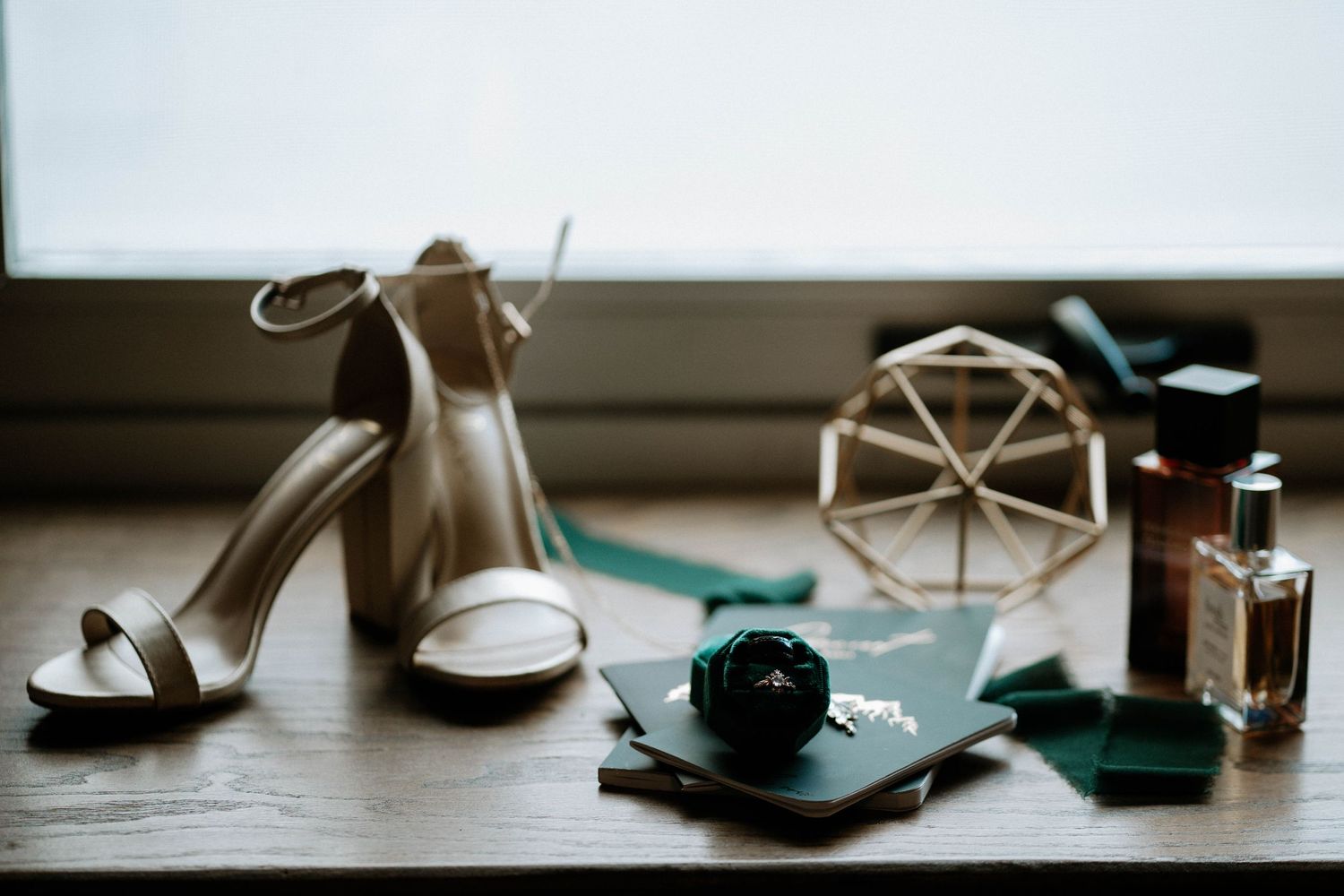 Bridal flat lay with nude heeled sandals, green accessories, perfume, and geometric decor on a wooden surface.