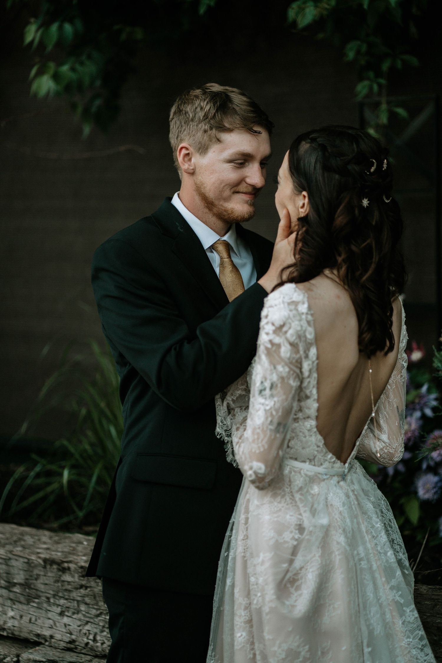Groom in black suit tenderly cups bride's face in lace wedding dress during romantic outdoor ceremony.