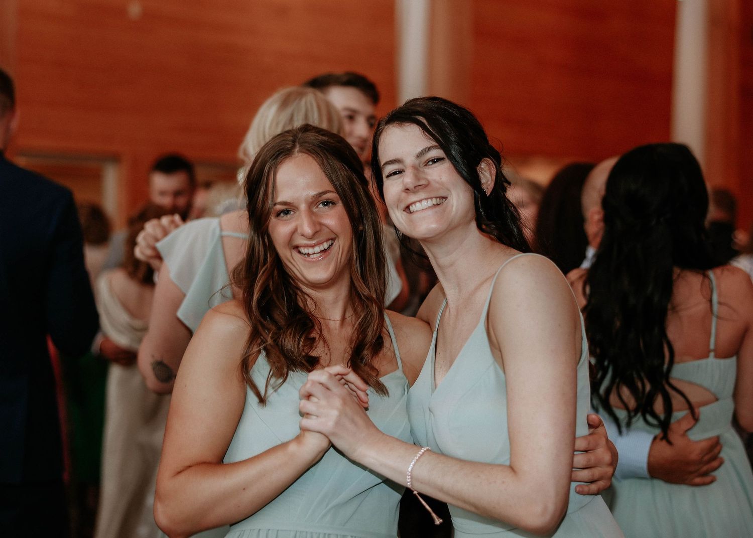 Two smiling bridesmaids in mint green dresses embrace and laugh together on the wedding dance floor.