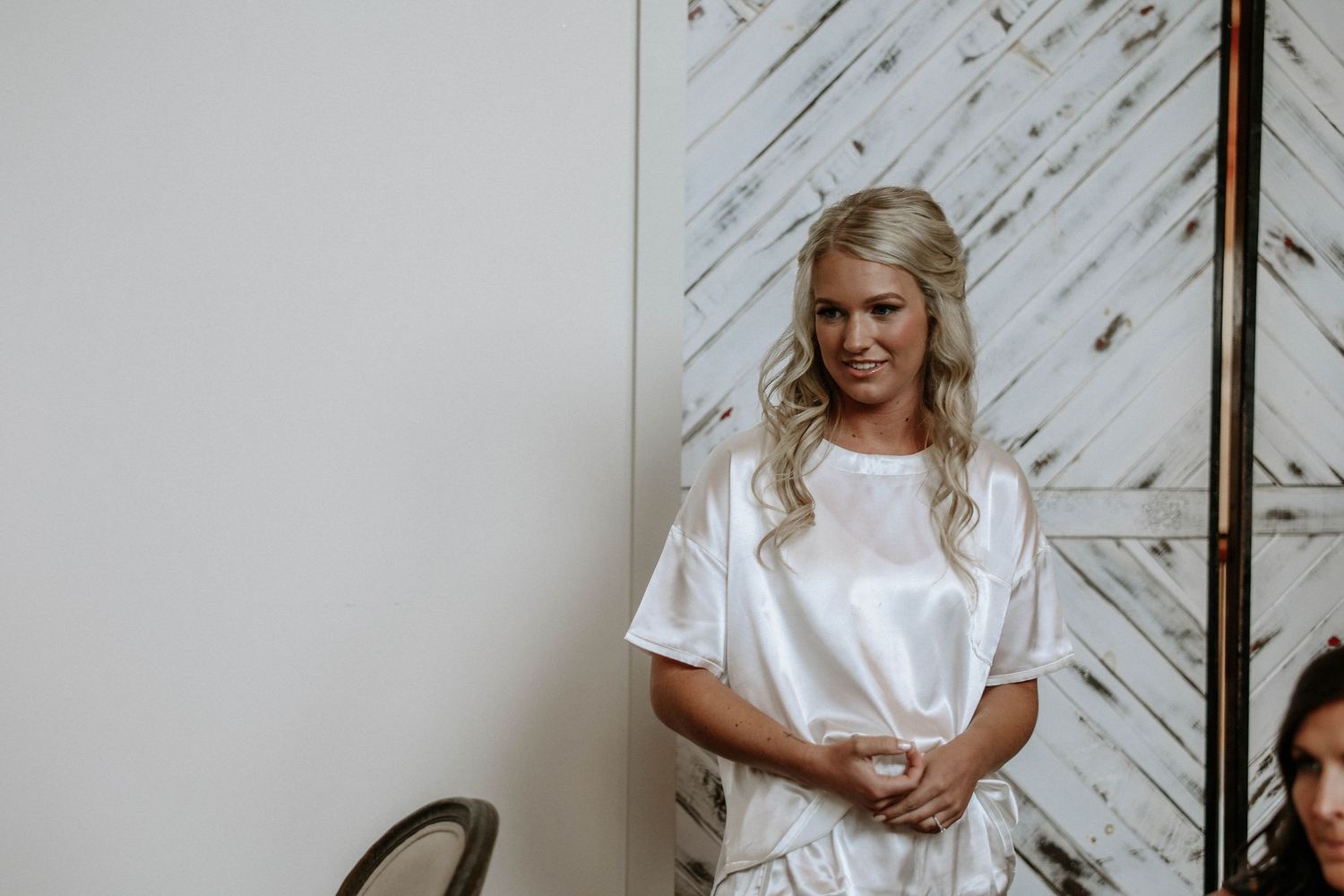 Blonde bride in white satin robe smiling during wedding preparations in a bright room with rustic barn doors.