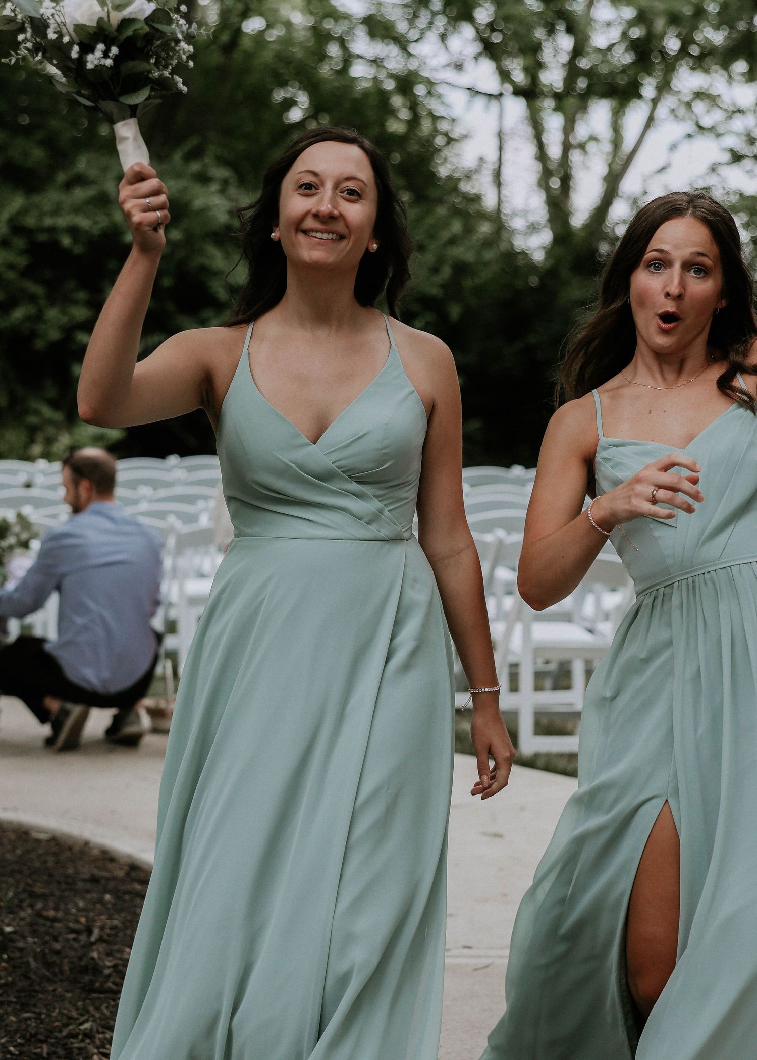 Two bridesmaids in sage green dresses walk down the aisle at an outdoor wedding ceremony.
