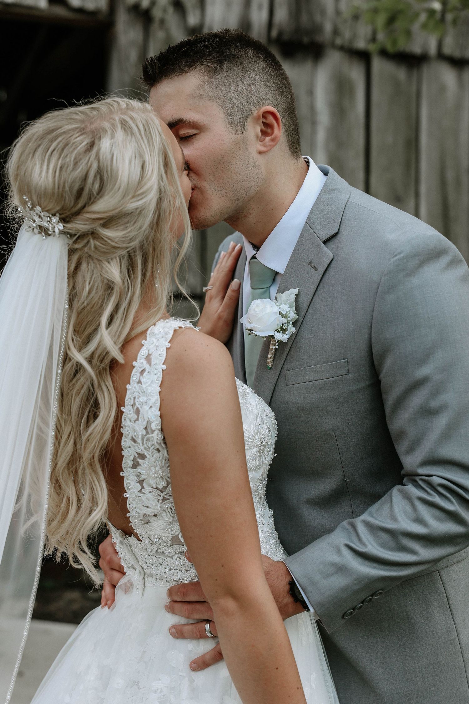 Bride and groom sharing a romantic kiss, bride in lace wedding dress with veil, groom in gray suit with white boutonniere.