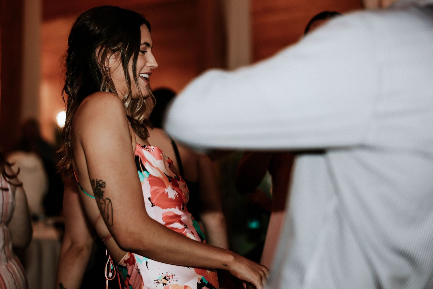 A woman in a floral dress laughs and dances at an indoor wedding reception celebration.