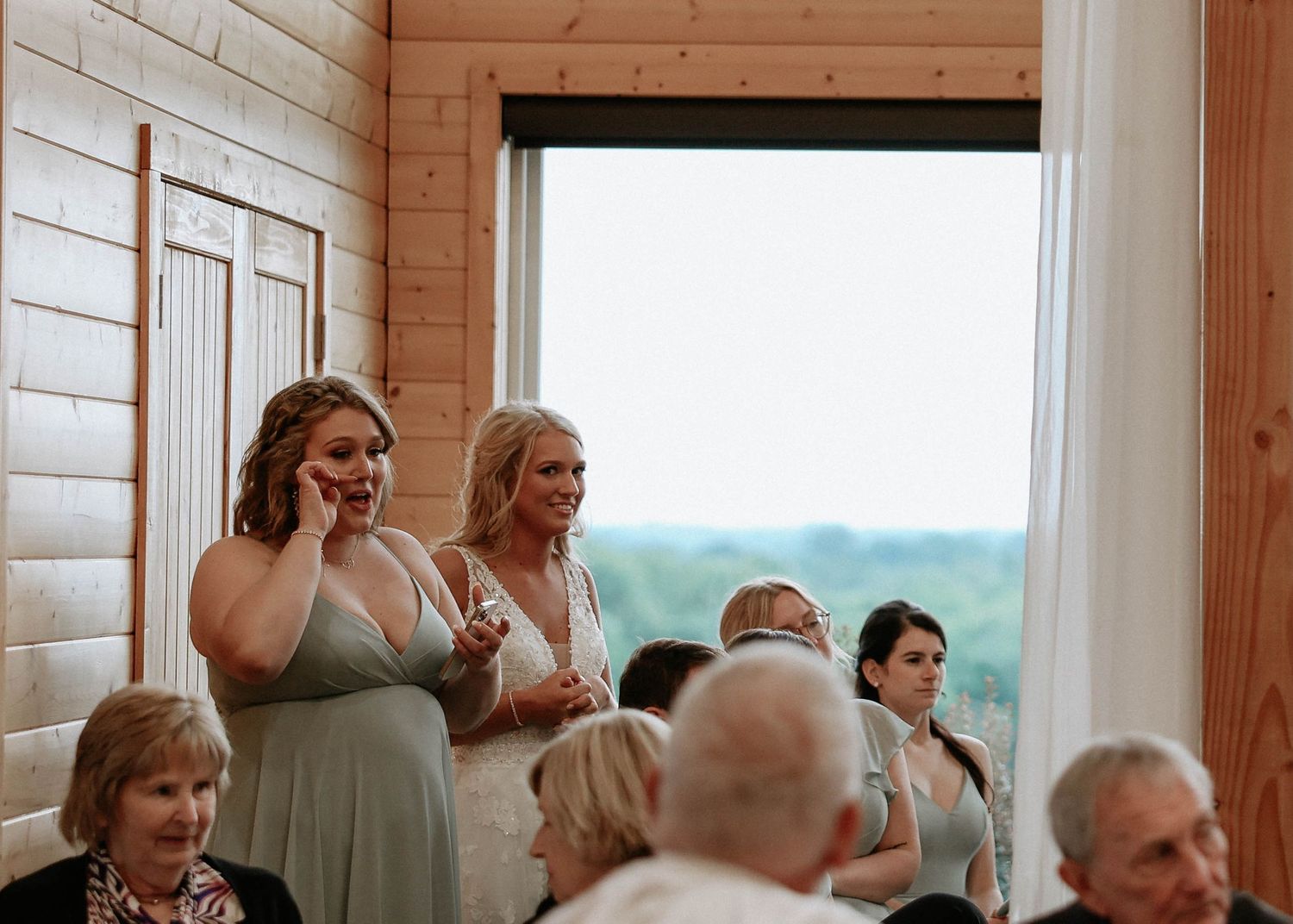 Emotional bridesmaids in sage dresses watch wedding ceremony at rustic barn venue.