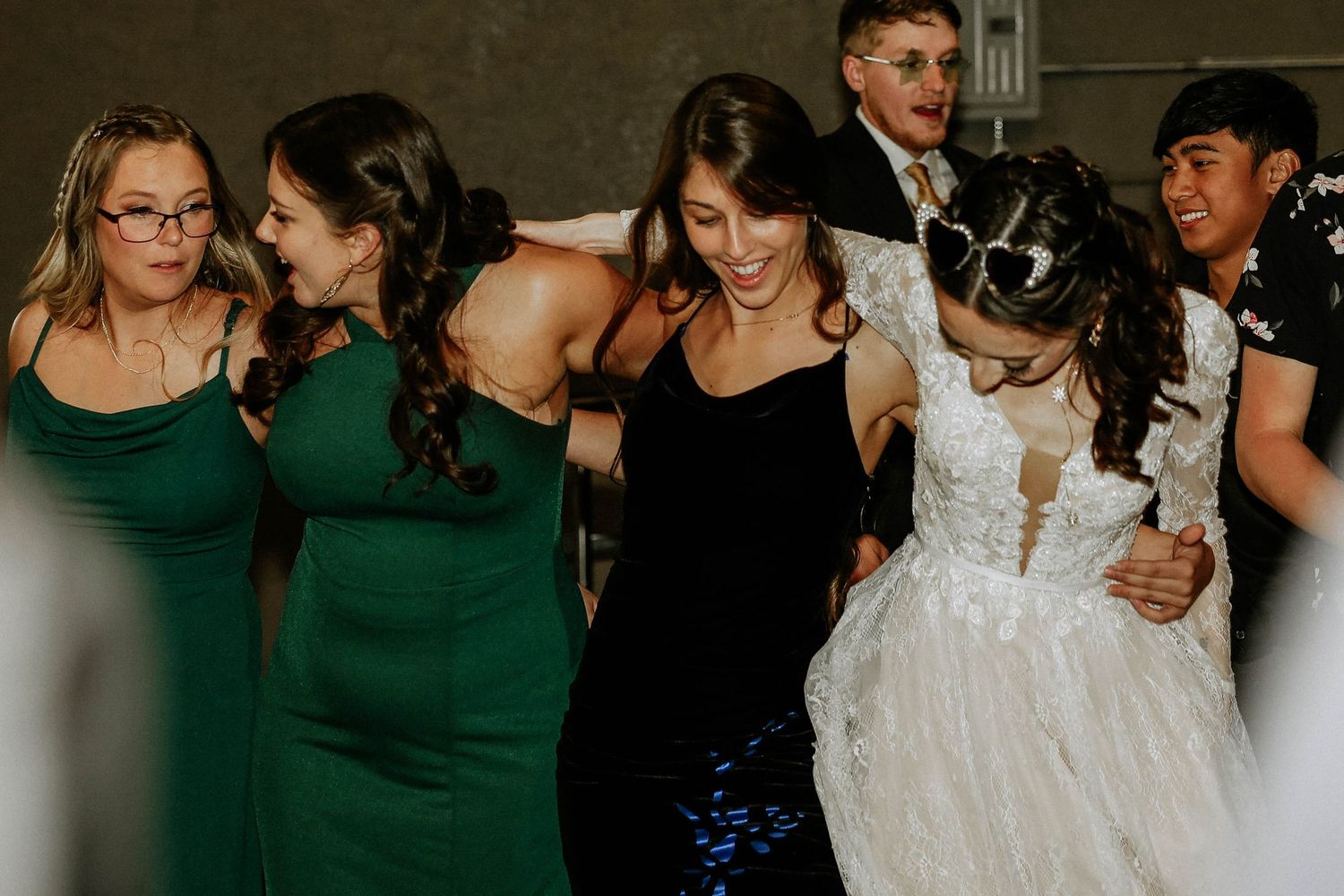 Bridesmaids in green dresses dancing joyfully with bride in white gown at wedding reception celebration.