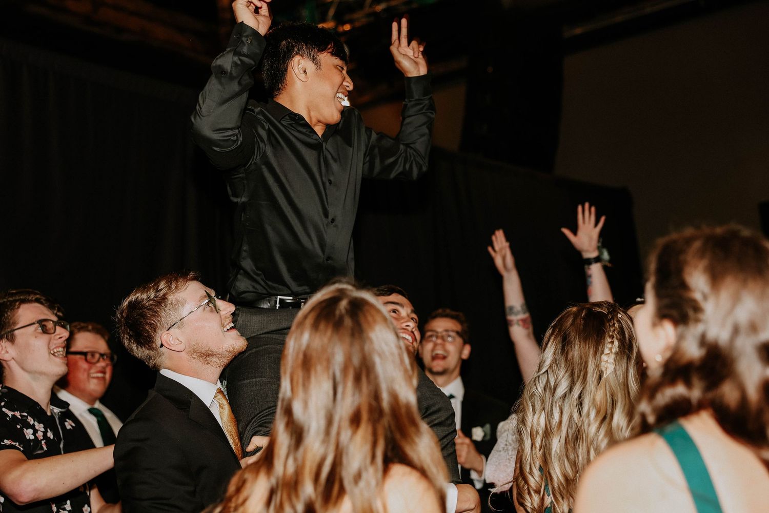Excited man stands above crowd at lively wedding reception dance floor, arms raised in celebration.