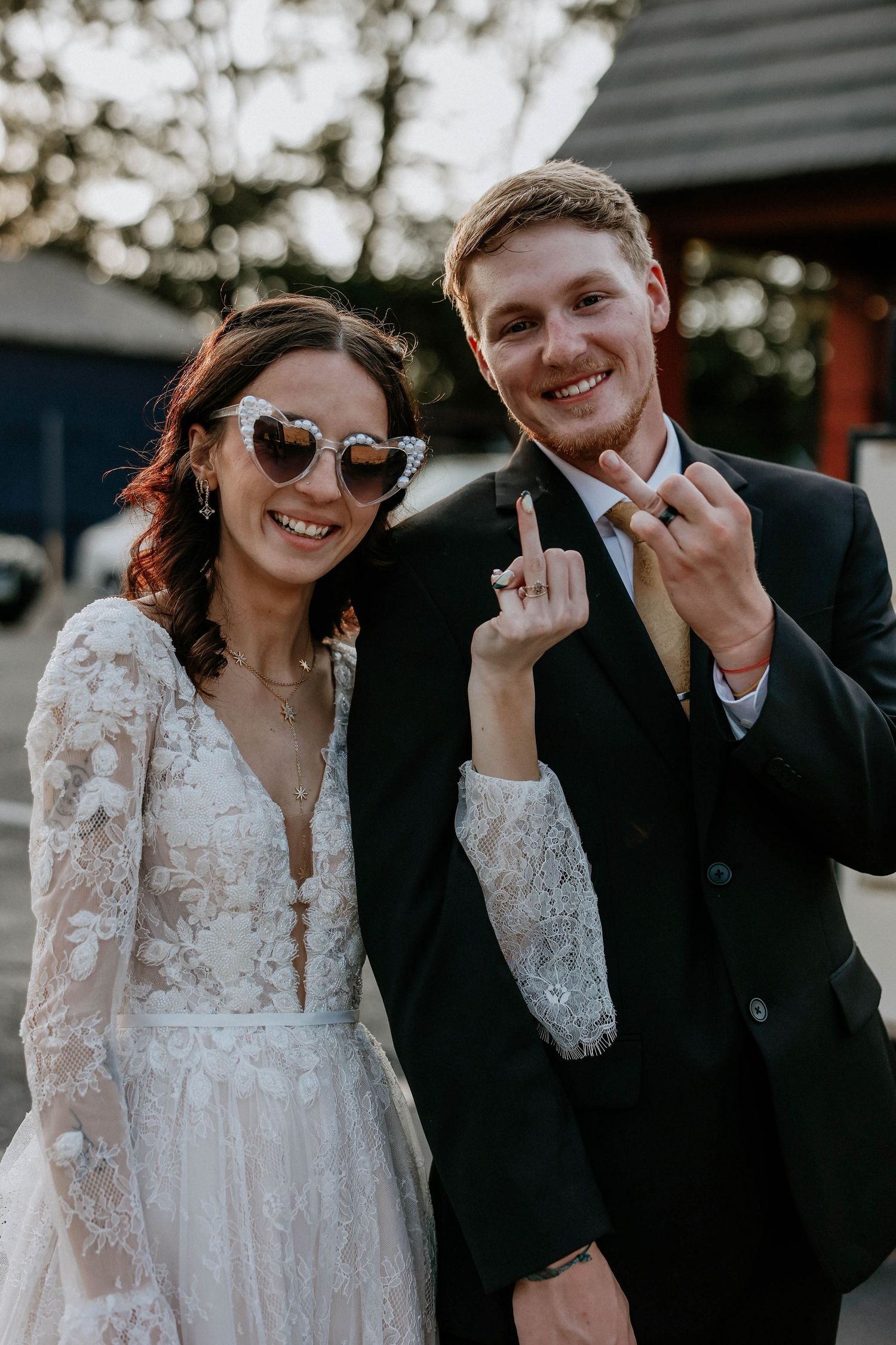 Smiling bride in lace gown with heart sunglasses and groom in black suit showing wedding rings outdoors.