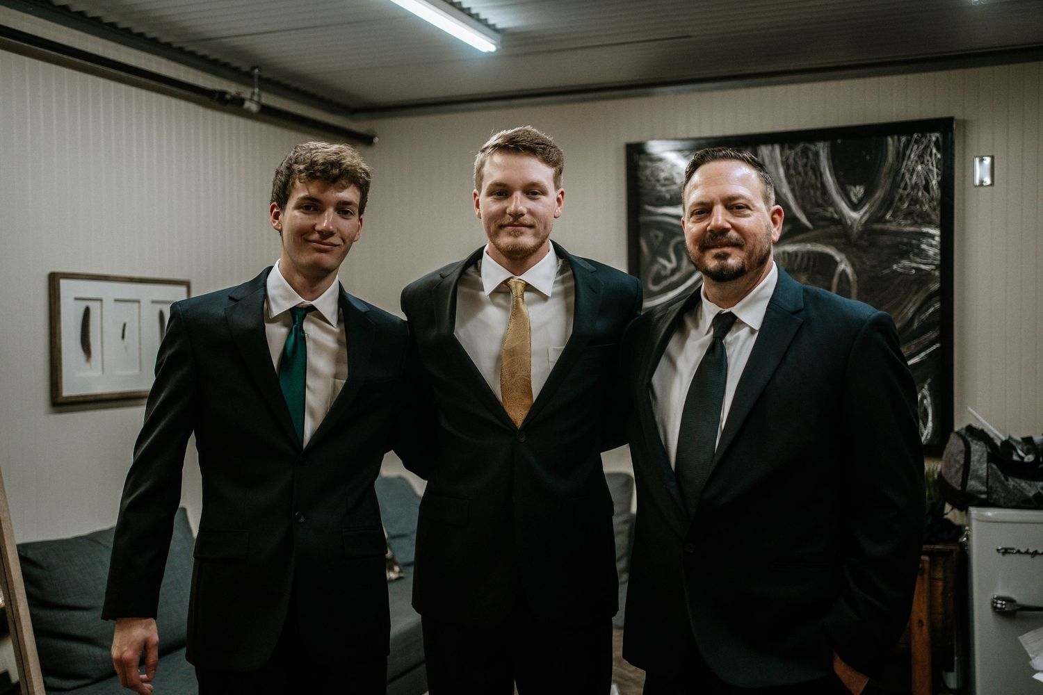 Three men in black suits and ties posing together smiling at a wedding celebration.