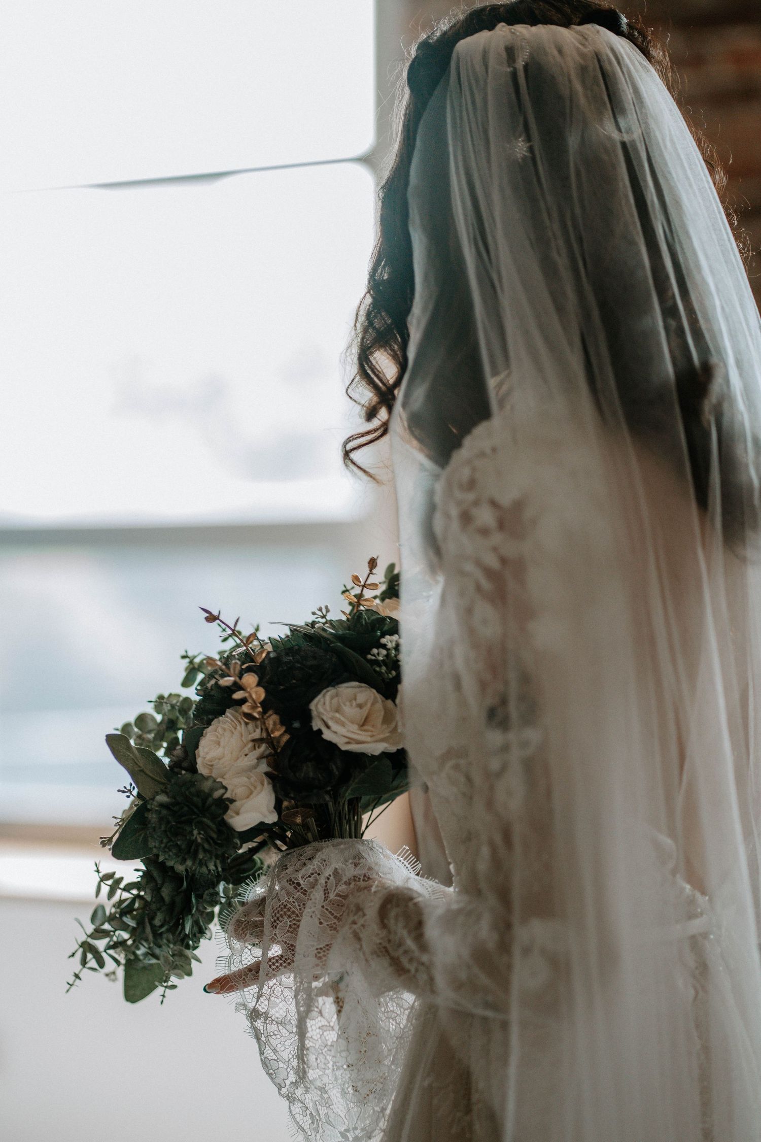 Bride in lace gown and veil holding a lush bouquet of white roses and greenery, standing by a window.