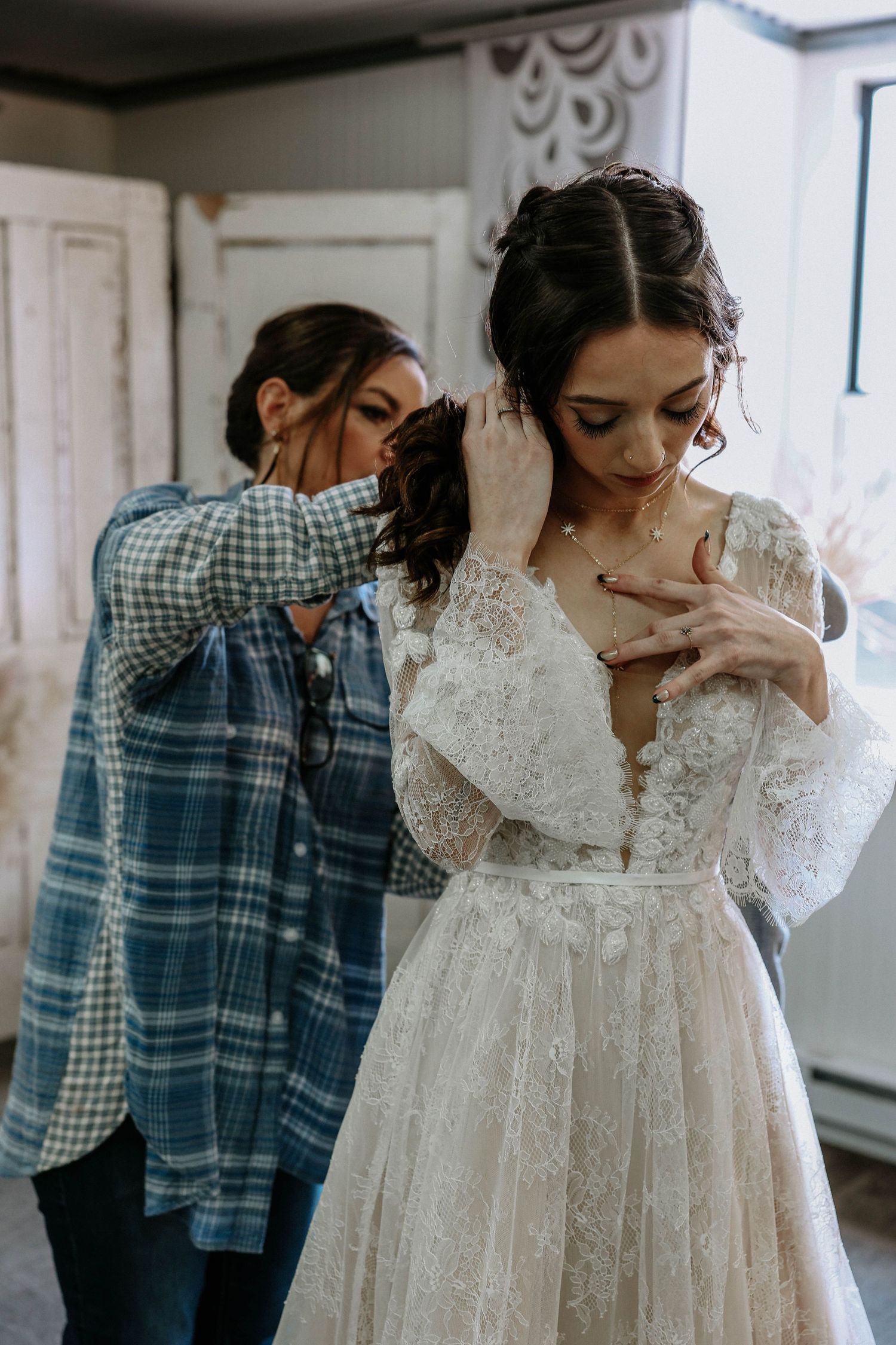 Bridesmaid in plaid shirt helps bride with earrings as she adjusts necklace in lace wedding gown.