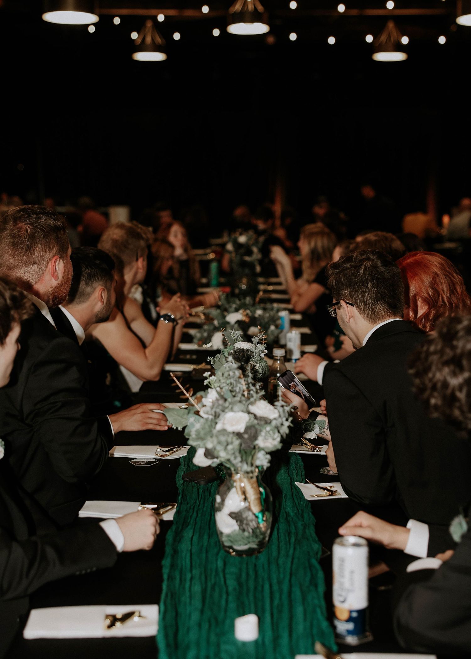 Elegant wedding reception with guests in formal attire seated at long tables adorned with green velvet runners and floral centerpieces.