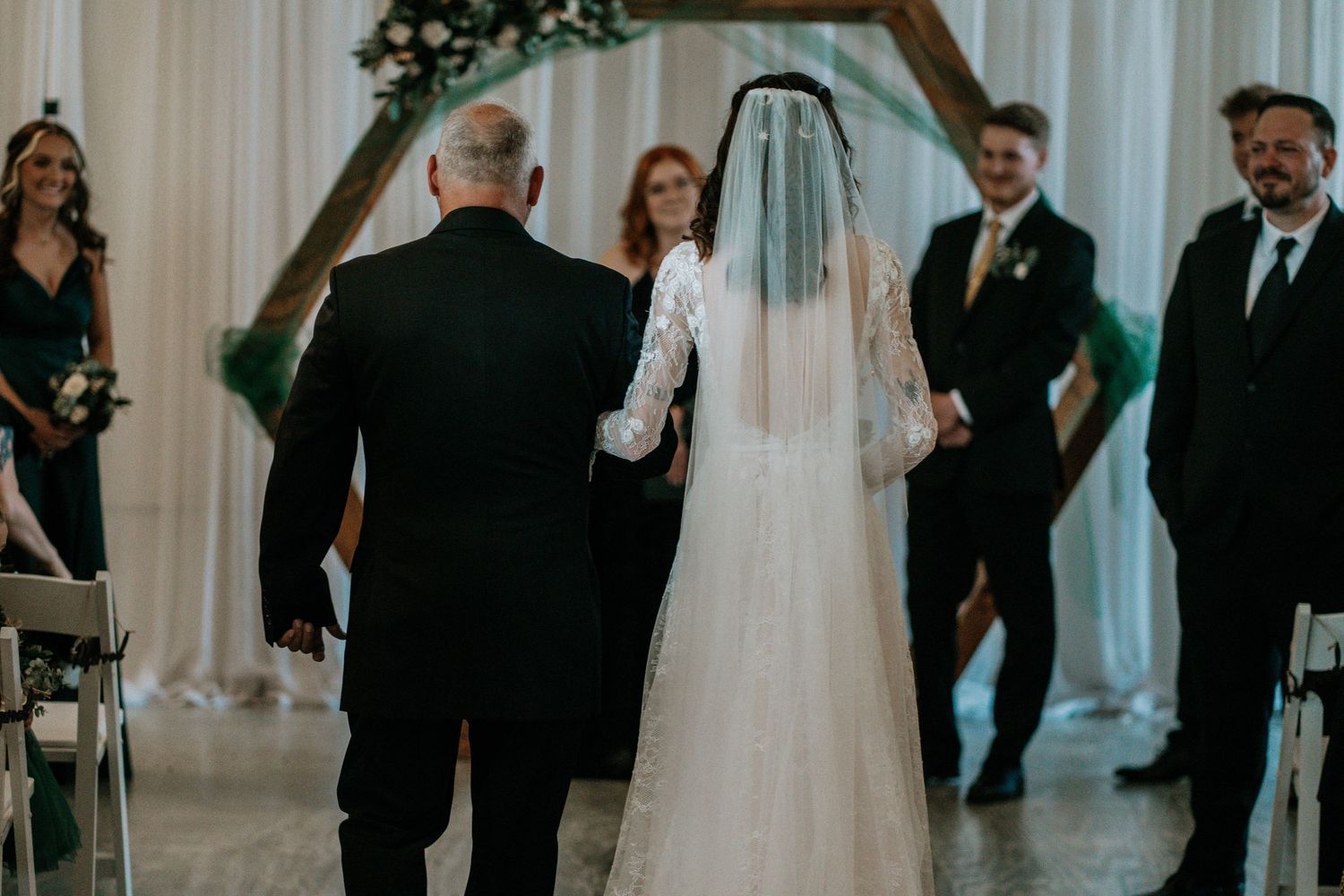 Bride in white lace gown and veil walks down the aisle with escort toward wooden arch ceremony backdrop.