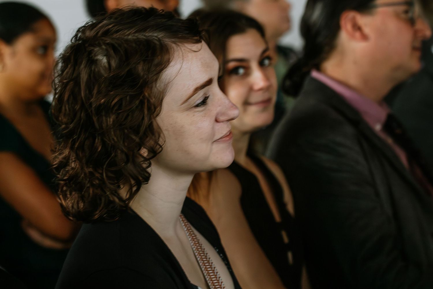 Young woman with curly hair smiling warmly at a wedding ceremony indoors.