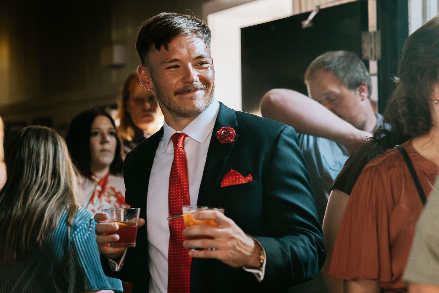 Dapper man in navy suit with red tie and pocket square holding cocktail at wedding.