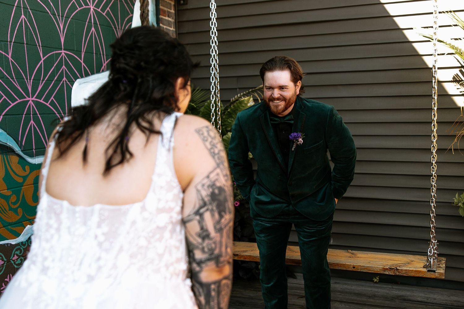 Groom in green velvet suit laughing with hands in pockets during first look with tattooed bride outdoors.