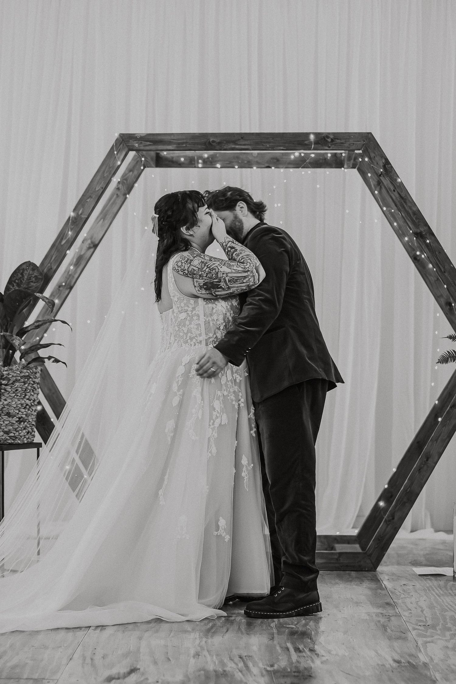 Black and white photo of newlyweds sharing first kiss under hexagonal wooden arch.