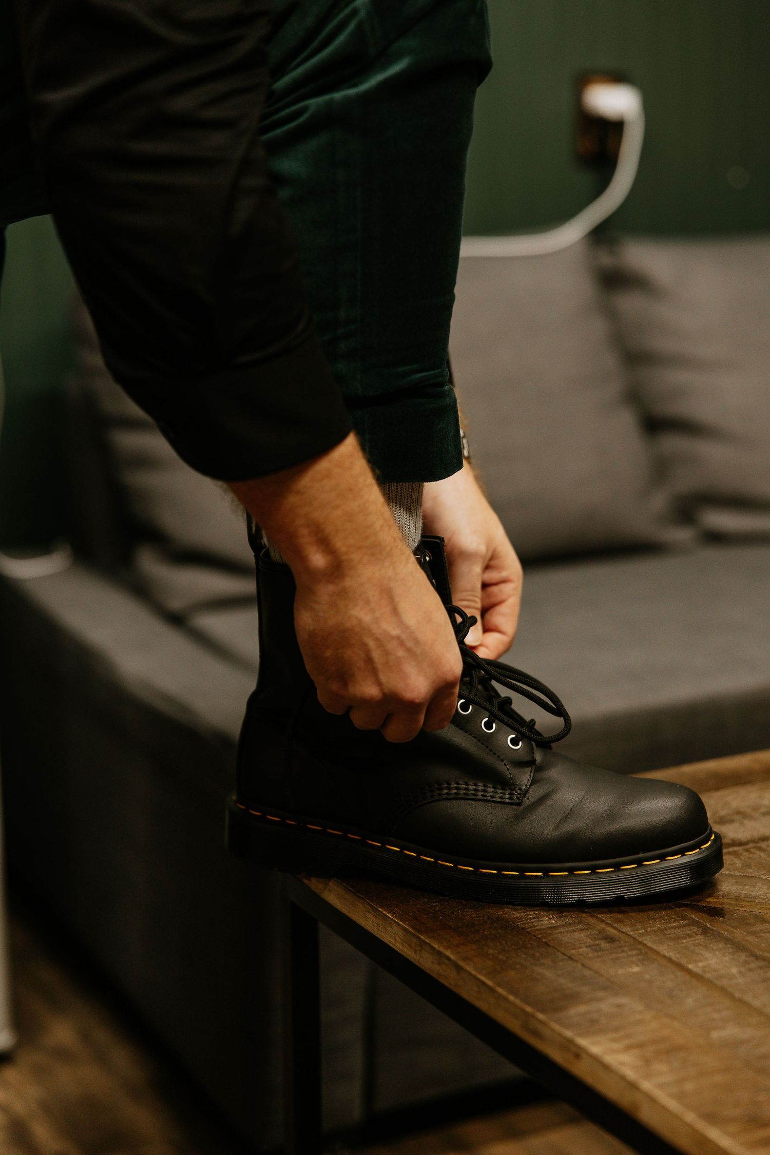 Man lacing up black Dr. Martens leather boots while seated near a gray sofa.
