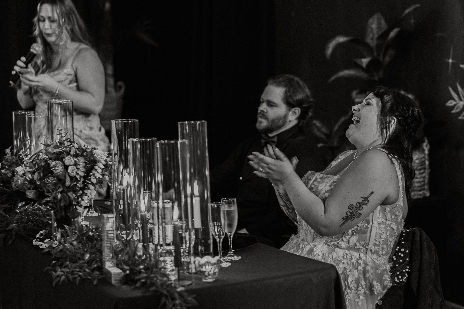 Black and white photo of wedding guests clapping during a speech at a candlelit reception table.