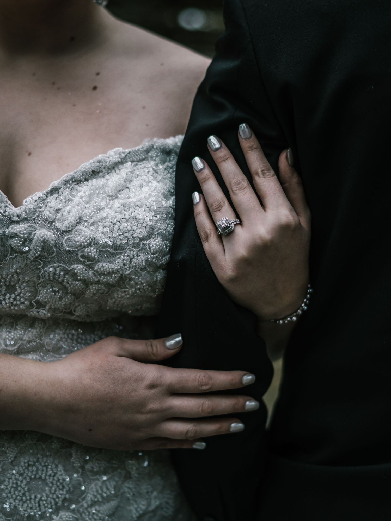 Close-up of bride's hand with diamond engagement ring and silver nails resting on groom's black tuxedo jacket.