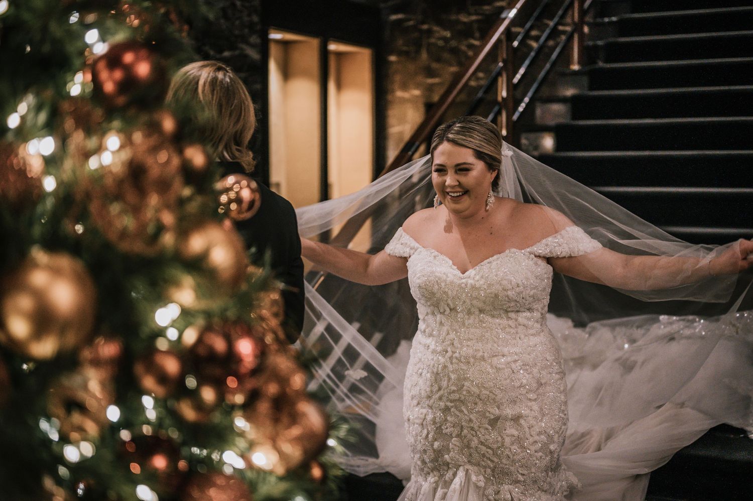 Bride in lace mermaid wedding gown with veil posing near decorated Christmas tree at venue.