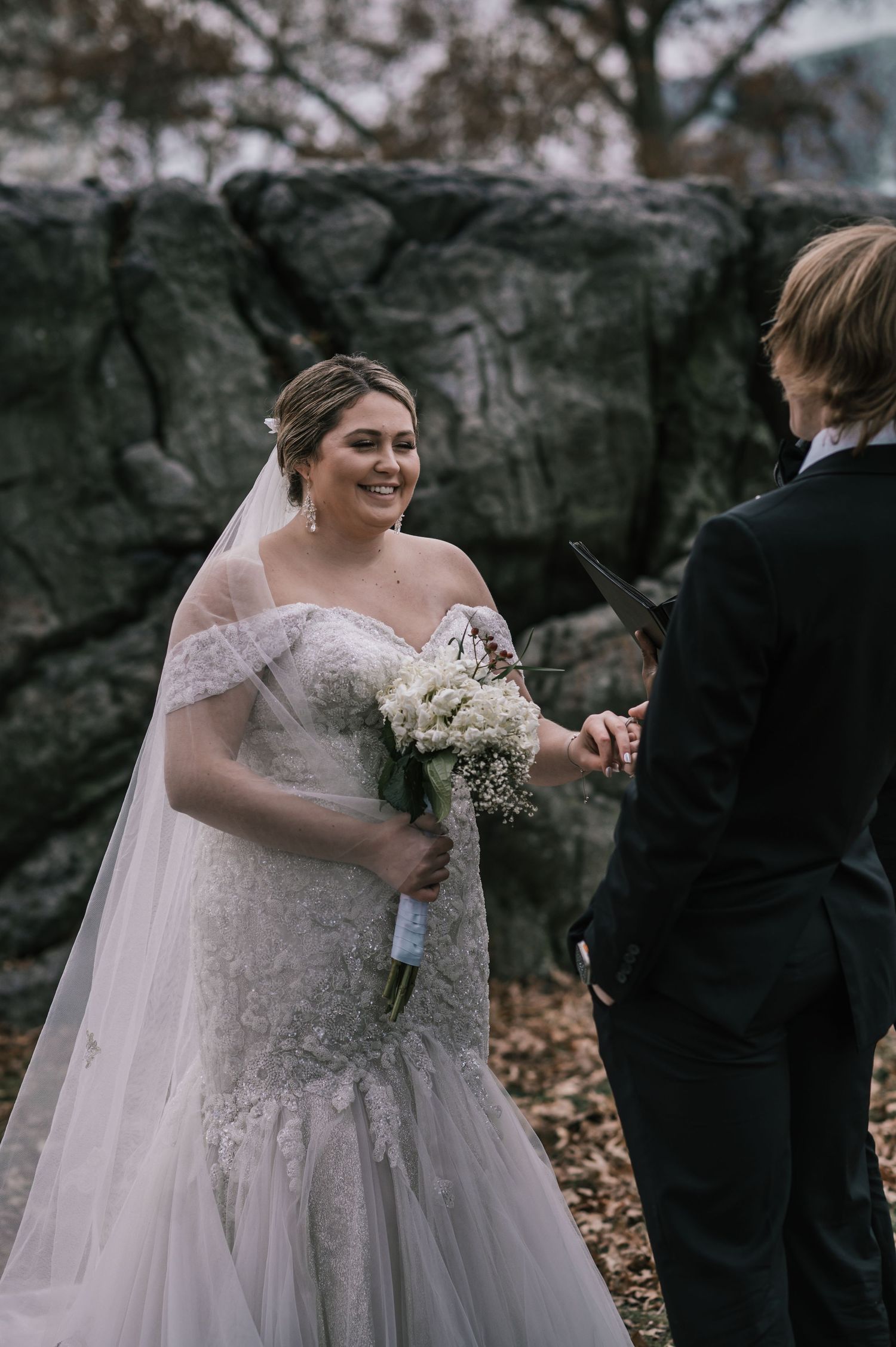 Radiant bride in lace mermaid gown holding white bouquet smiling during outdoor wedding ceremony by rocks.