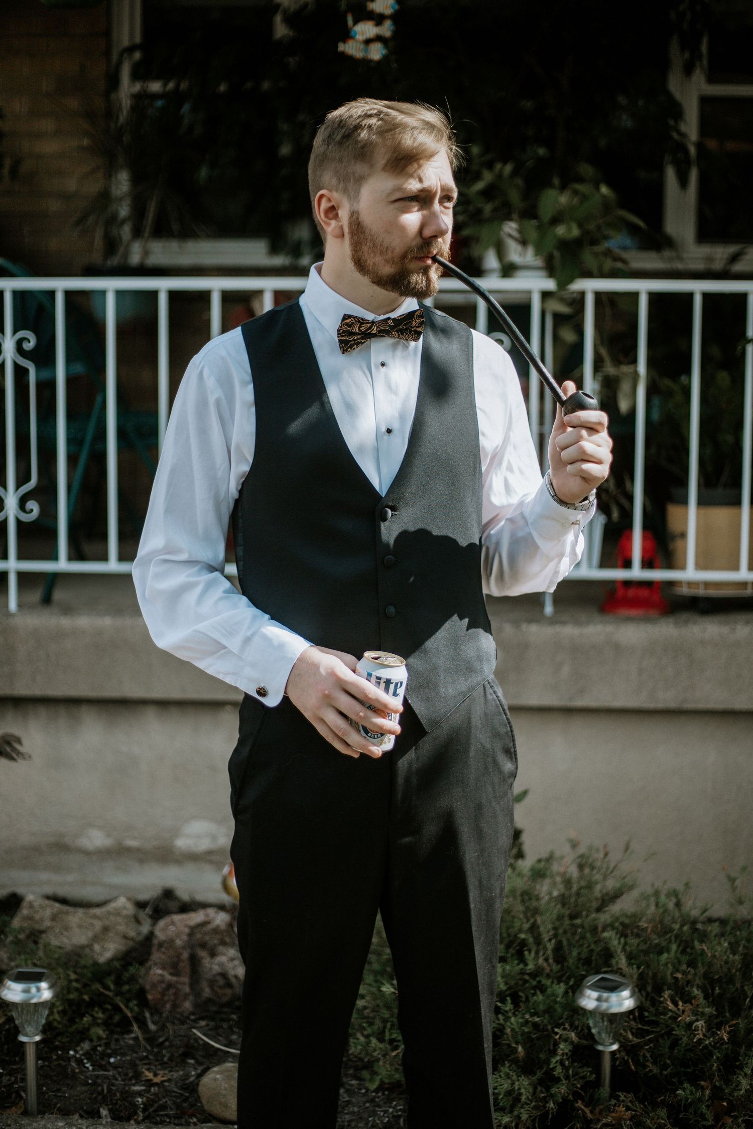 Dapper groom in black vest and bow tie smokes a pipe while holding a beer can outdoors in Cincinnati, Ohio