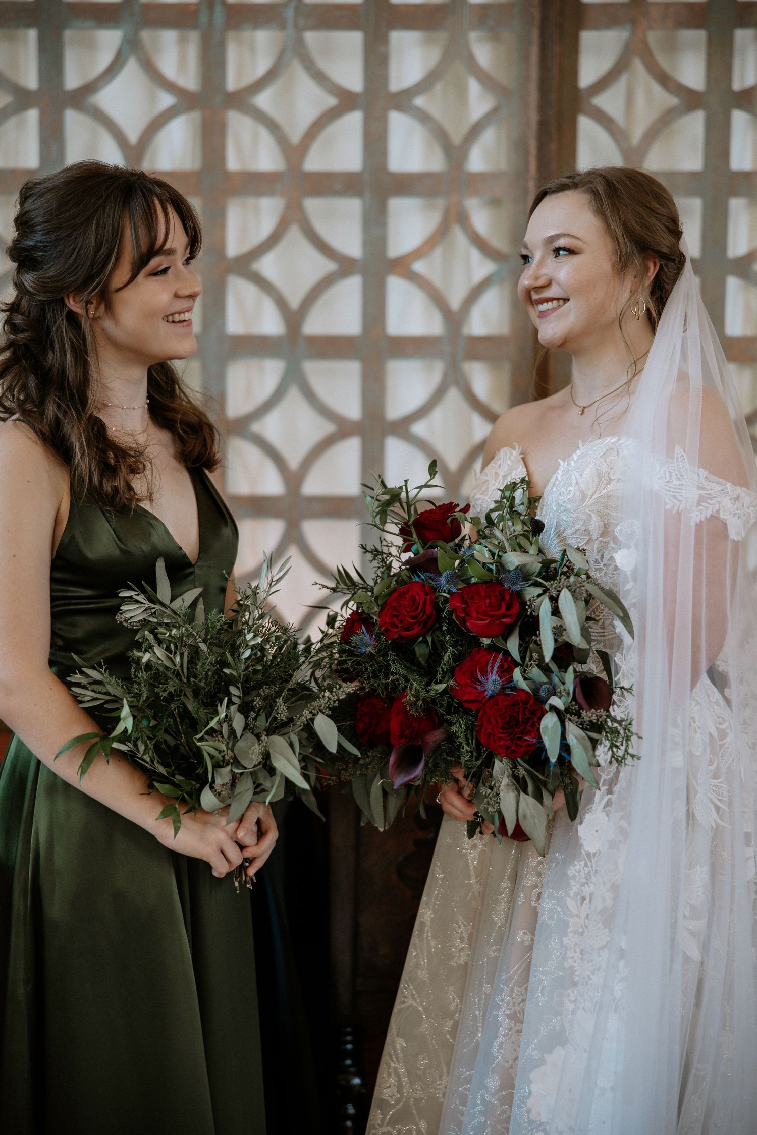 Bride in lace veil gown and bridesmaid in olive green dress smile while holding floral bouquets at Urban Artifact in Cincinnati, Ohio