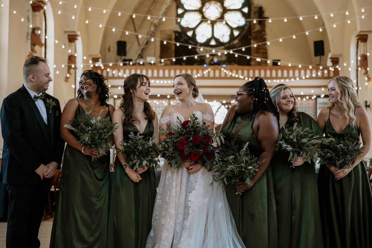 Laughing bride in lace gown poses with bridesmaids in olive green dresses holding red bouquets at Urban Artifact in Cincinnati, Ohio
