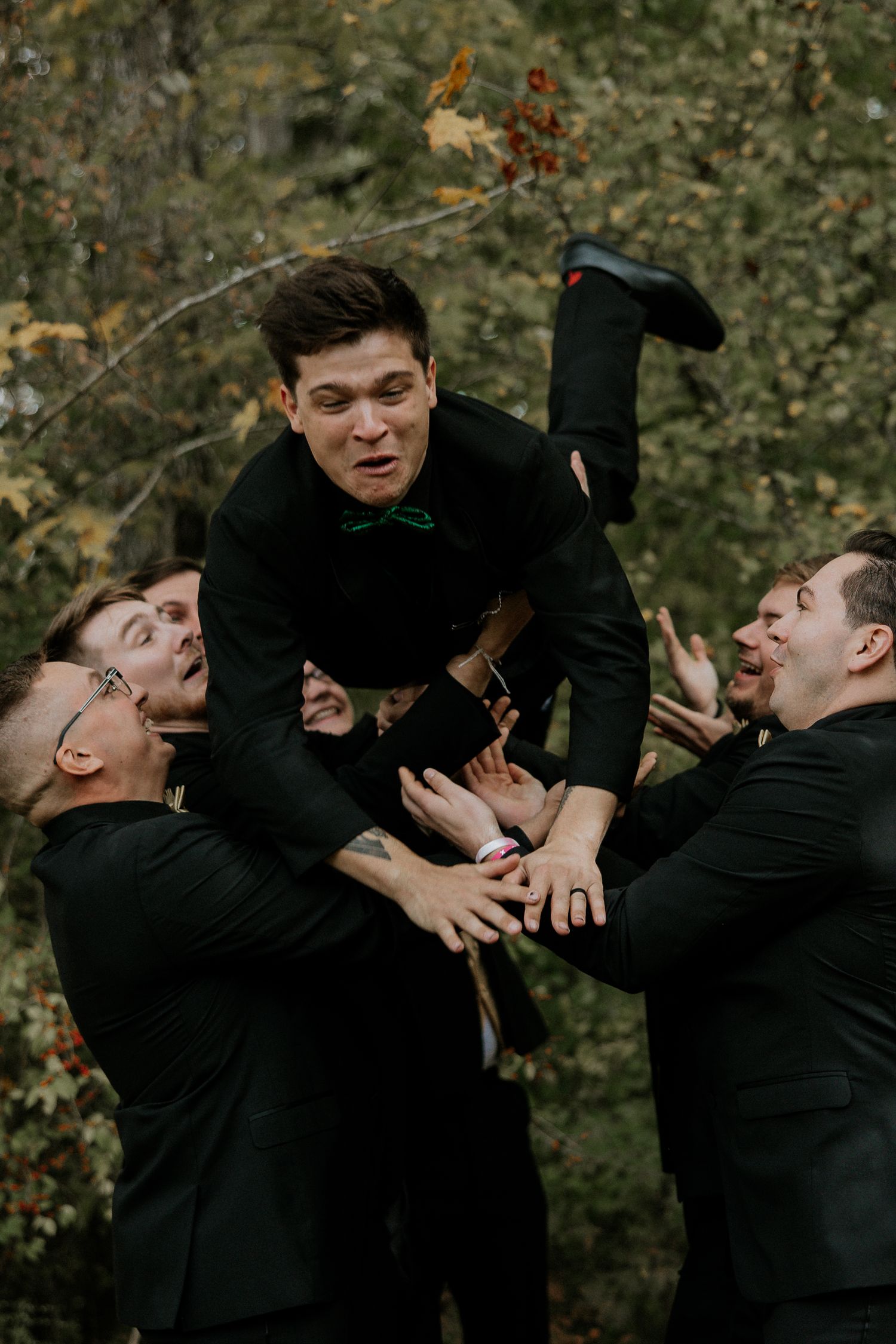 Groomsmen in black suits lifting the groom in the air outdoors among autumn trees at The Barn at Wolf Creek in Akron, Ohio