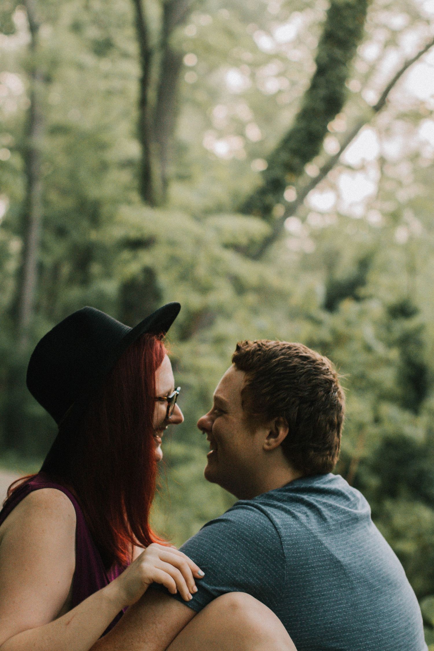Happy couple laughing together outdoors in lush green forest, woman with red hair wearing black hat in Cincinnati, Ohio.