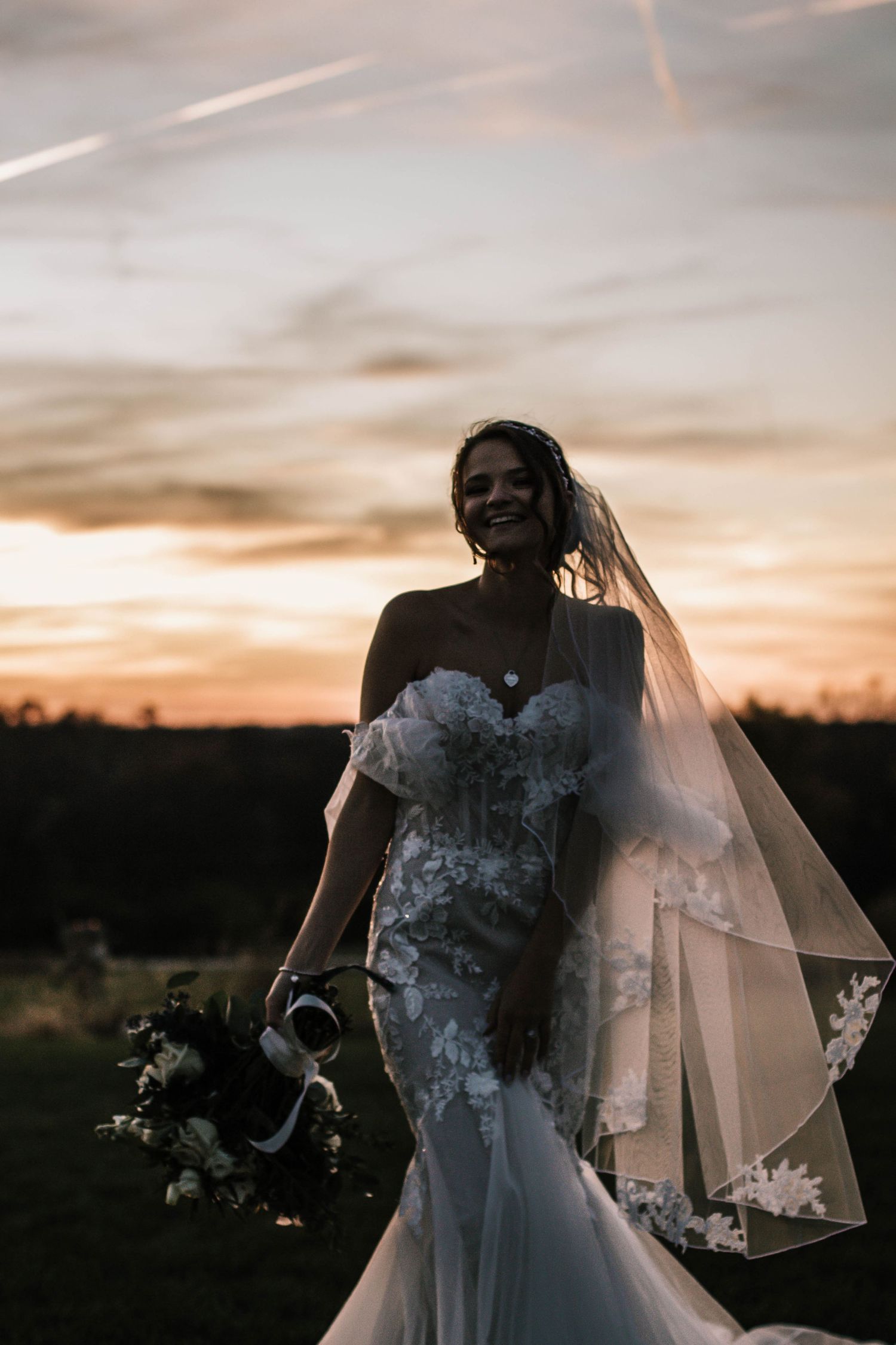 Radiant bride in lace mermaid gown and veil smiling at golden sunset holding bouquet outdoors at Stone Valley Meadows in Dayton, Ohio
