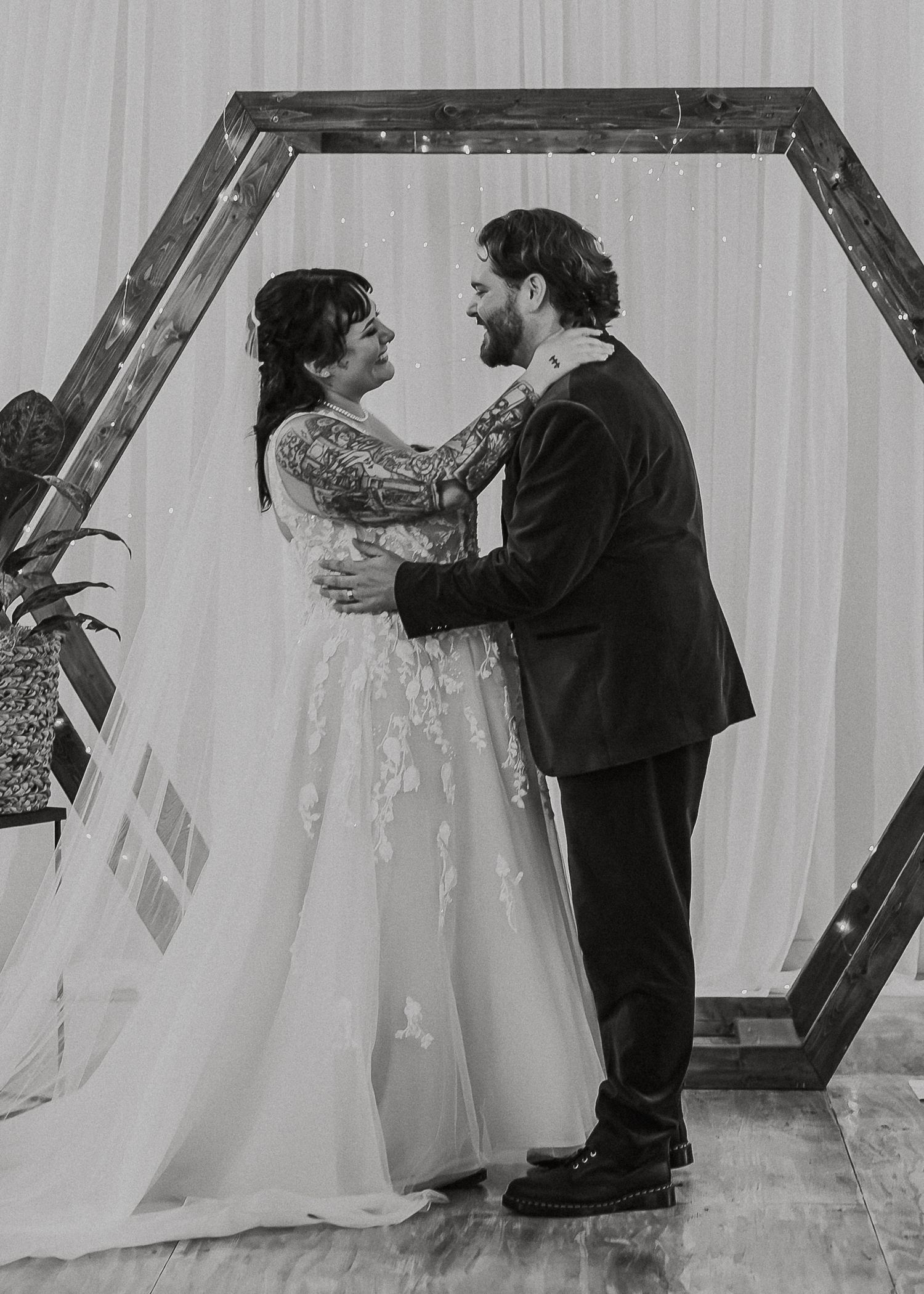 Black and white photo of newlyweds embracing and smiling under hexagonal wooden arch.