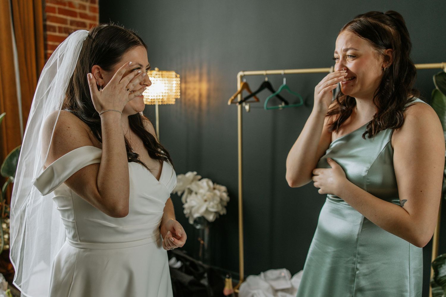 Bride in white gown and bridesmaid in sage green share an emotional laughing moment before the wedding.