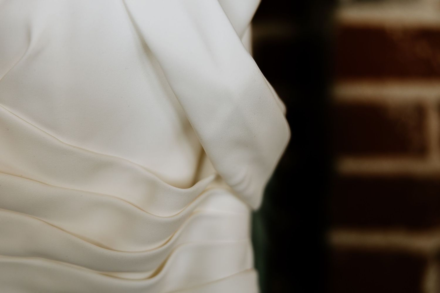 Close-up detail of elegant ivory satin wedding dress with draped fabric against exposed brick wall.