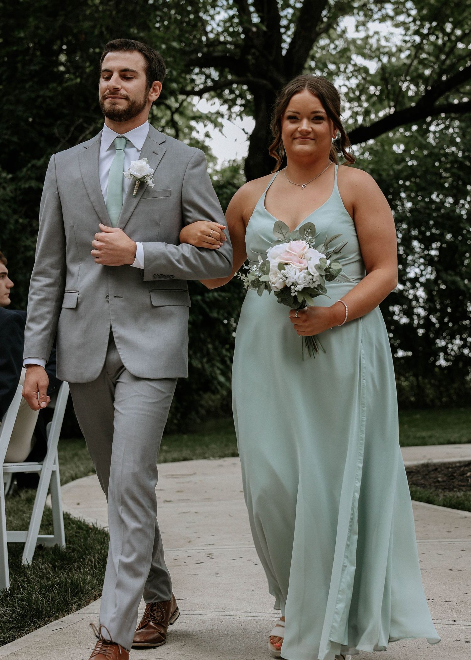 Groomsman in gray suit escorting bridesmaid in sage green gown down the outdoor wedding aisle.