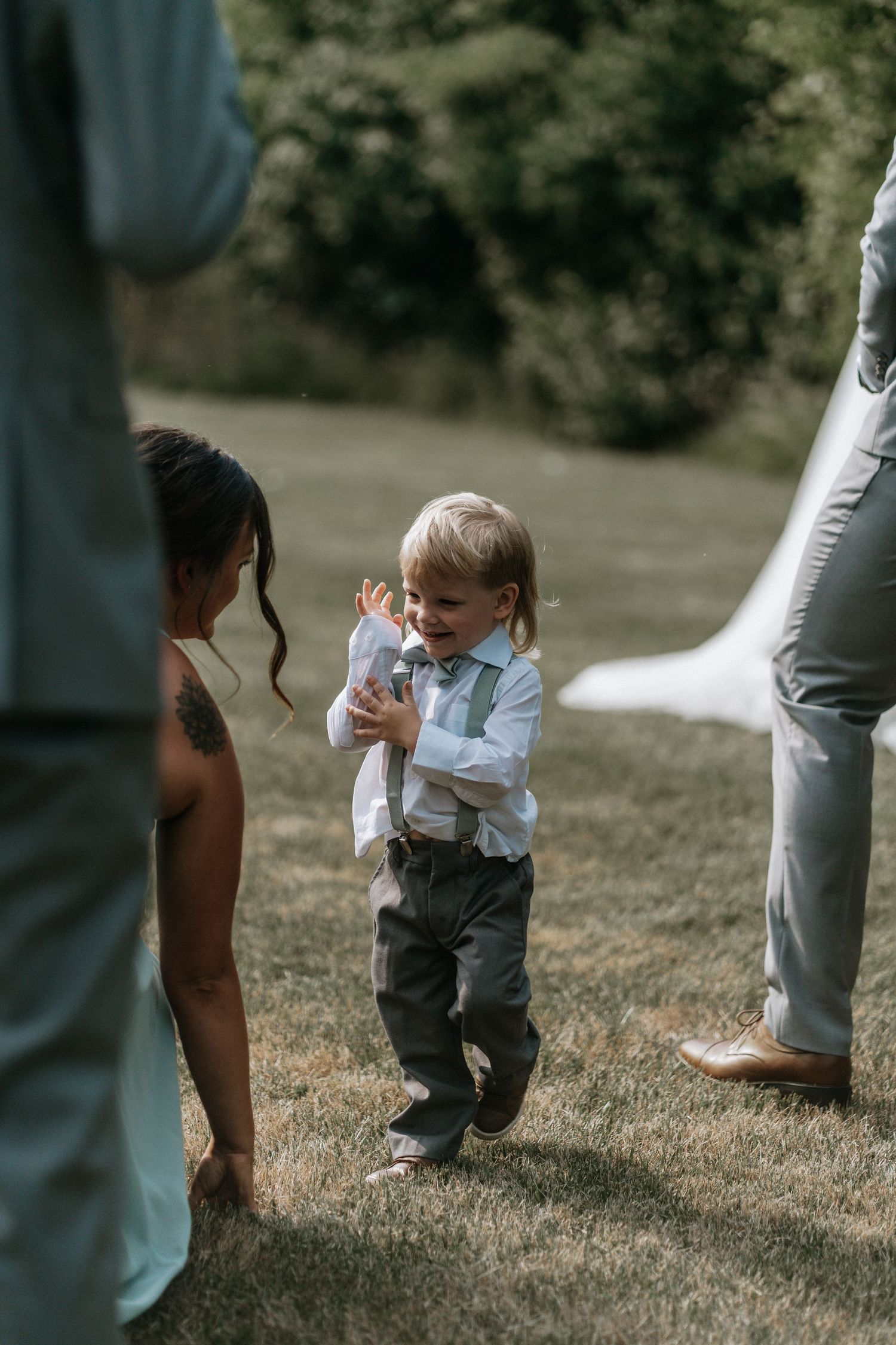 Adorable ring bearer in white shirt and suspenders laughing during an outdoor wedding ceremony on green grass.