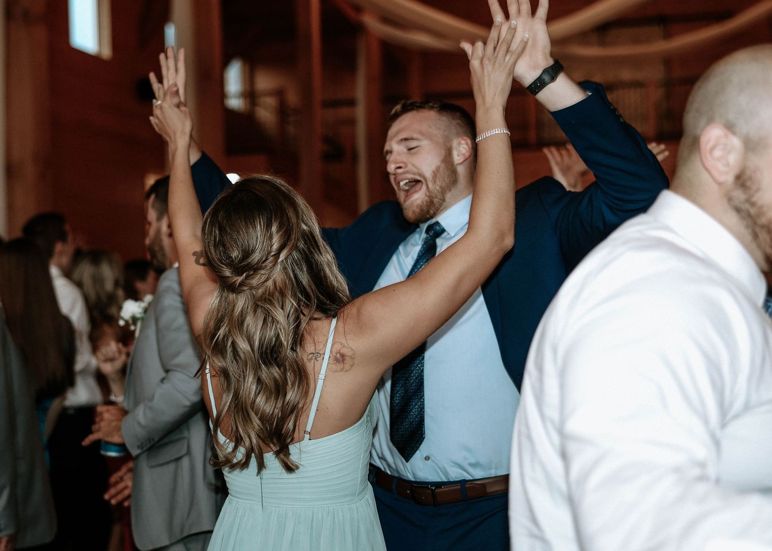 Guests dancing joyfully at a wedding reception in a rustic barn venue, with hands raised in celebration.