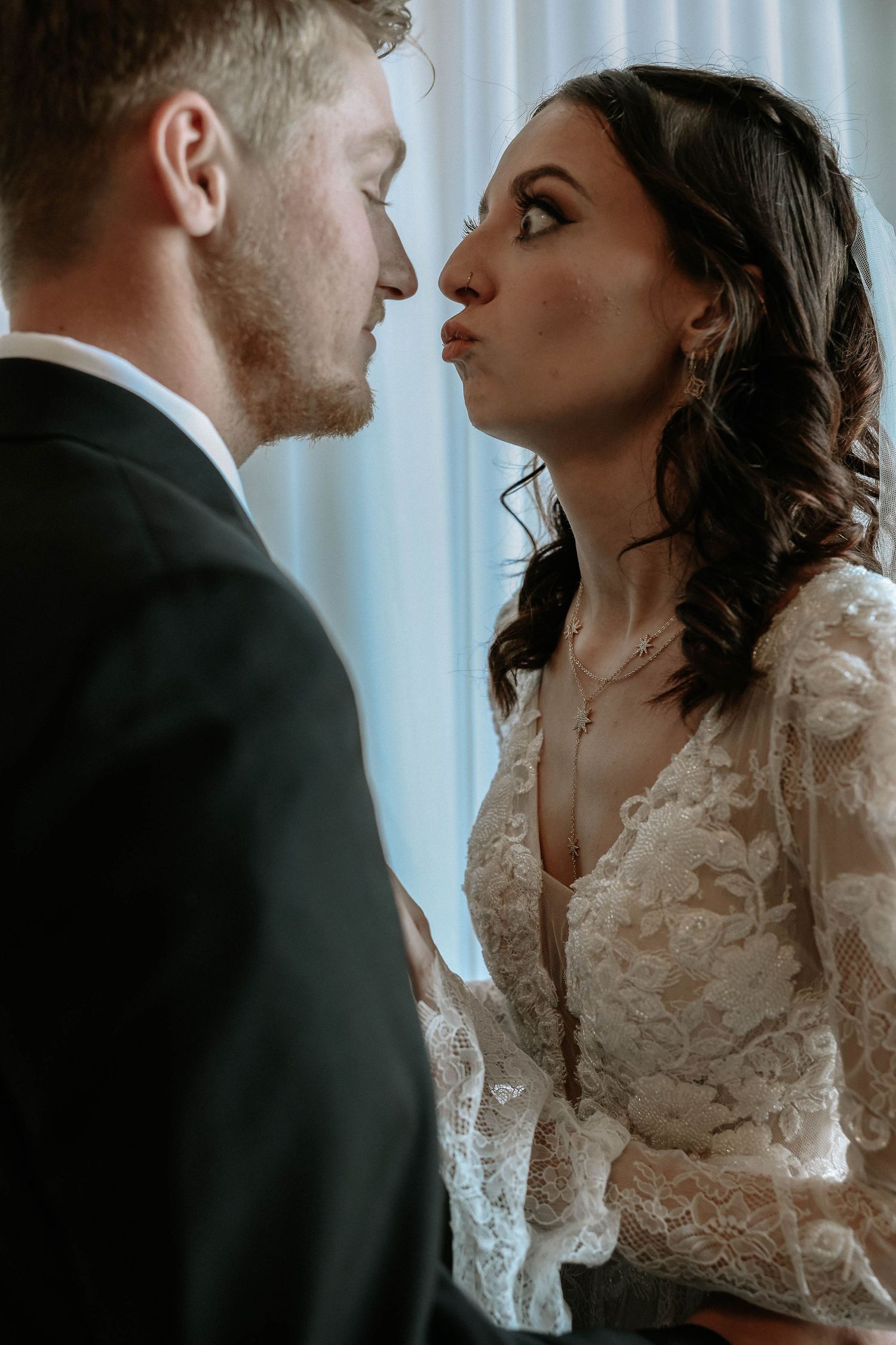 Bride in lace wedding dress puckers lips for a kiss with groom in black suit during romantic wedding moment.