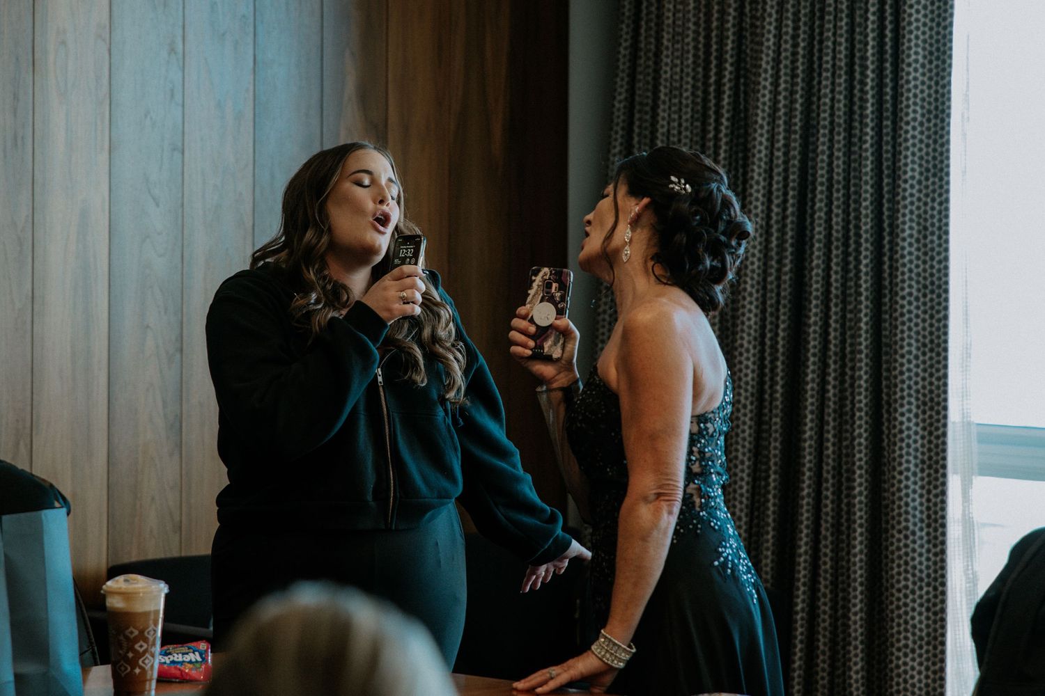 Two women in elegant dark dresses sipping drinks and laughing together at a formal event.