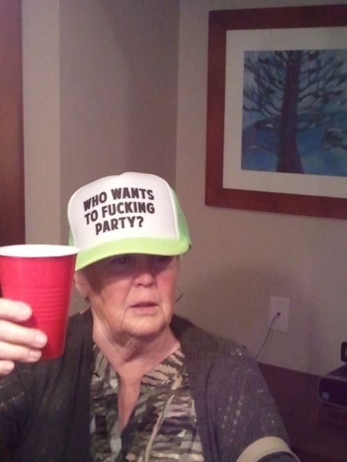Elderly woman holding red Solo cup wearing neon green trucker hat that reads Who Wants To Fucking Party.